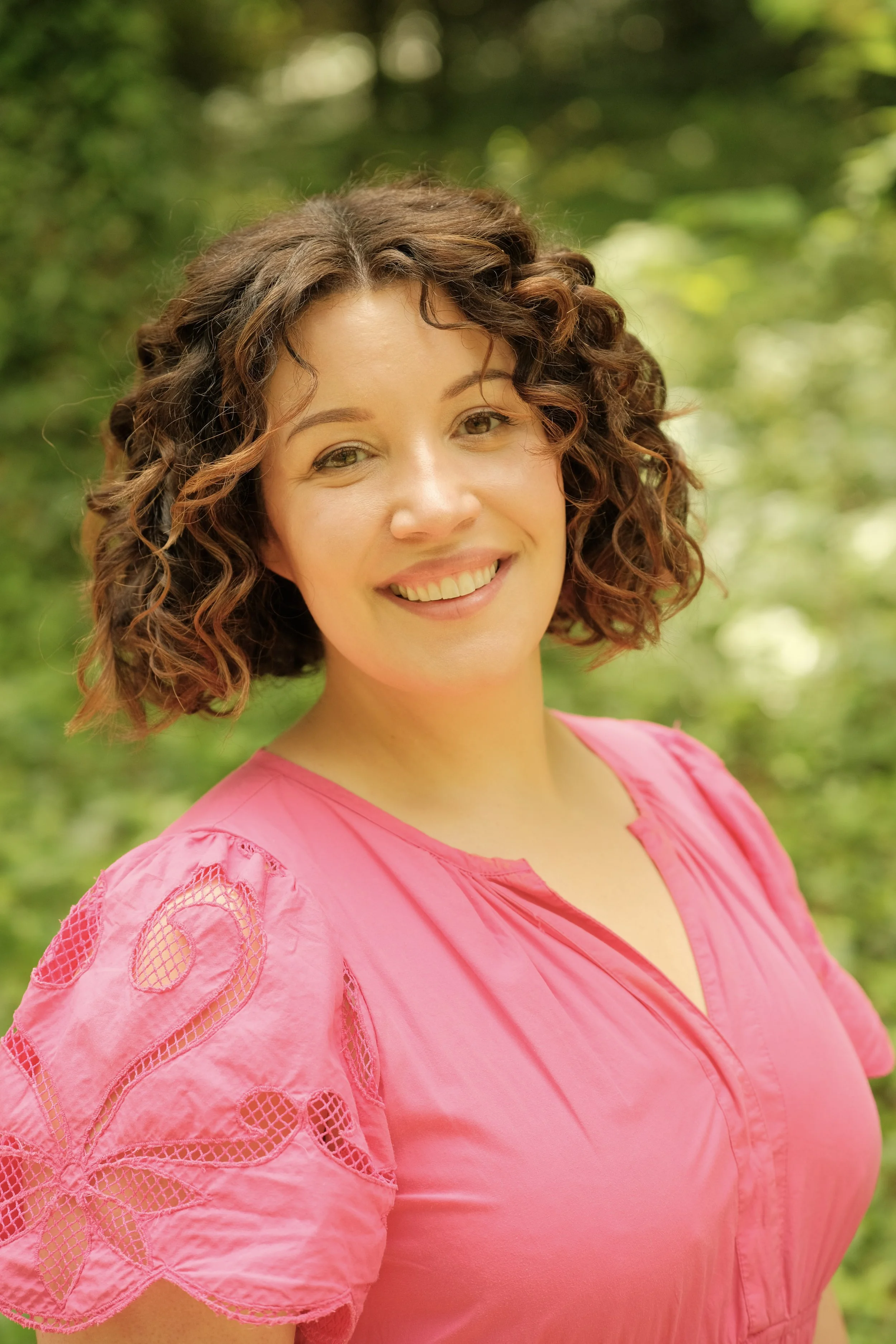 A woman with curly brown hair wearing a pink top with lace and embroidery details, smiling outdoors with green foliage in the background.