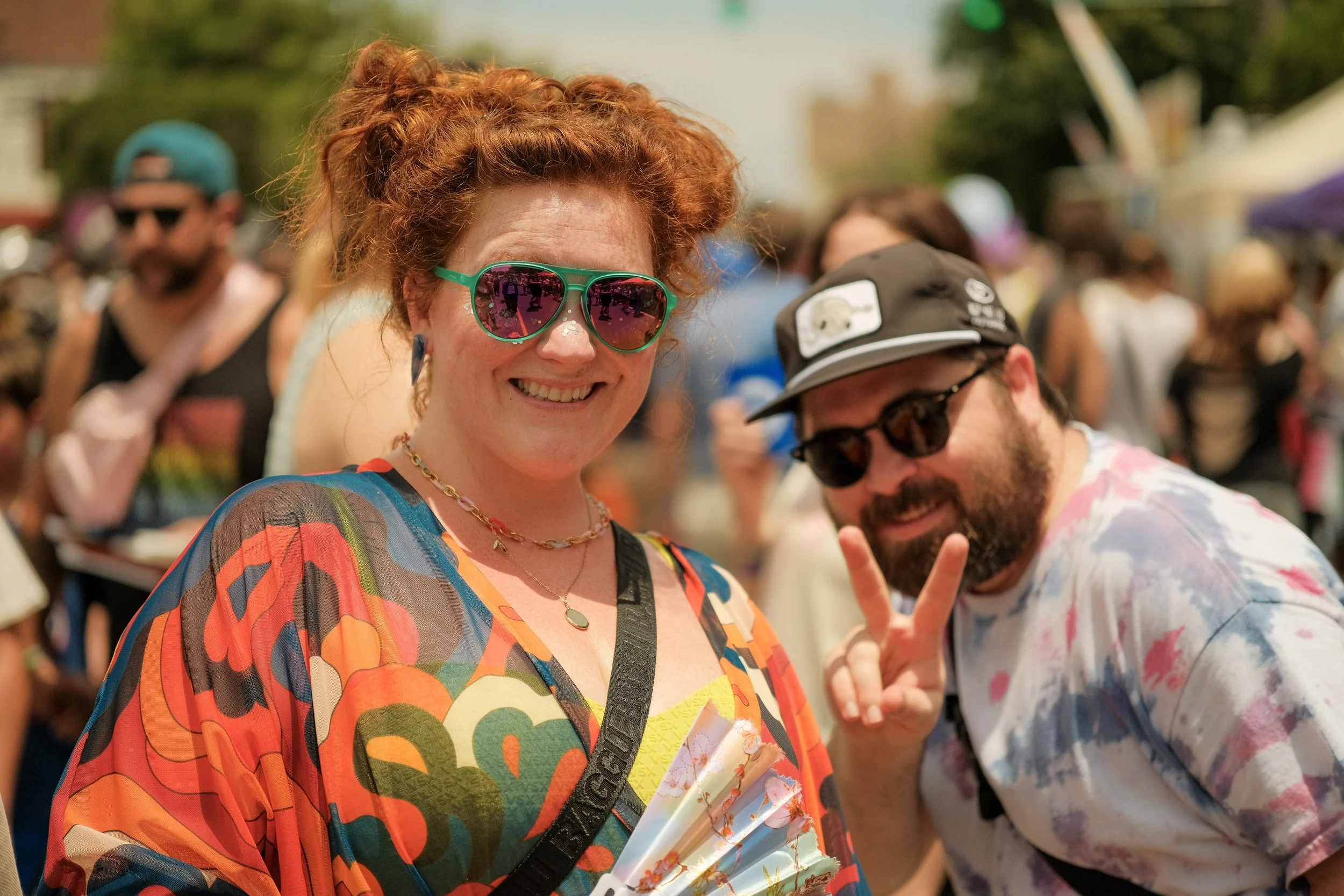 A woman with curly red hair wearing colorful sunglasses and a multicolored patterned dress smiling at a crowded outdoor event.