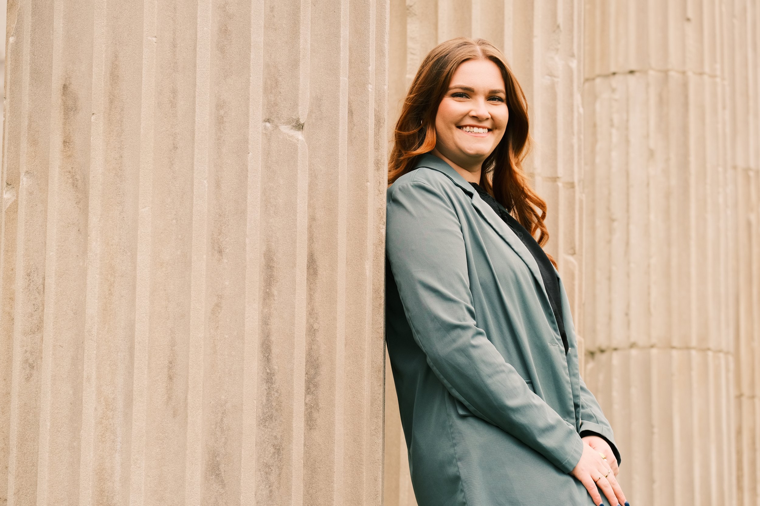 A woman with long red hair smiling, leaning against a large stone column, outdoors.