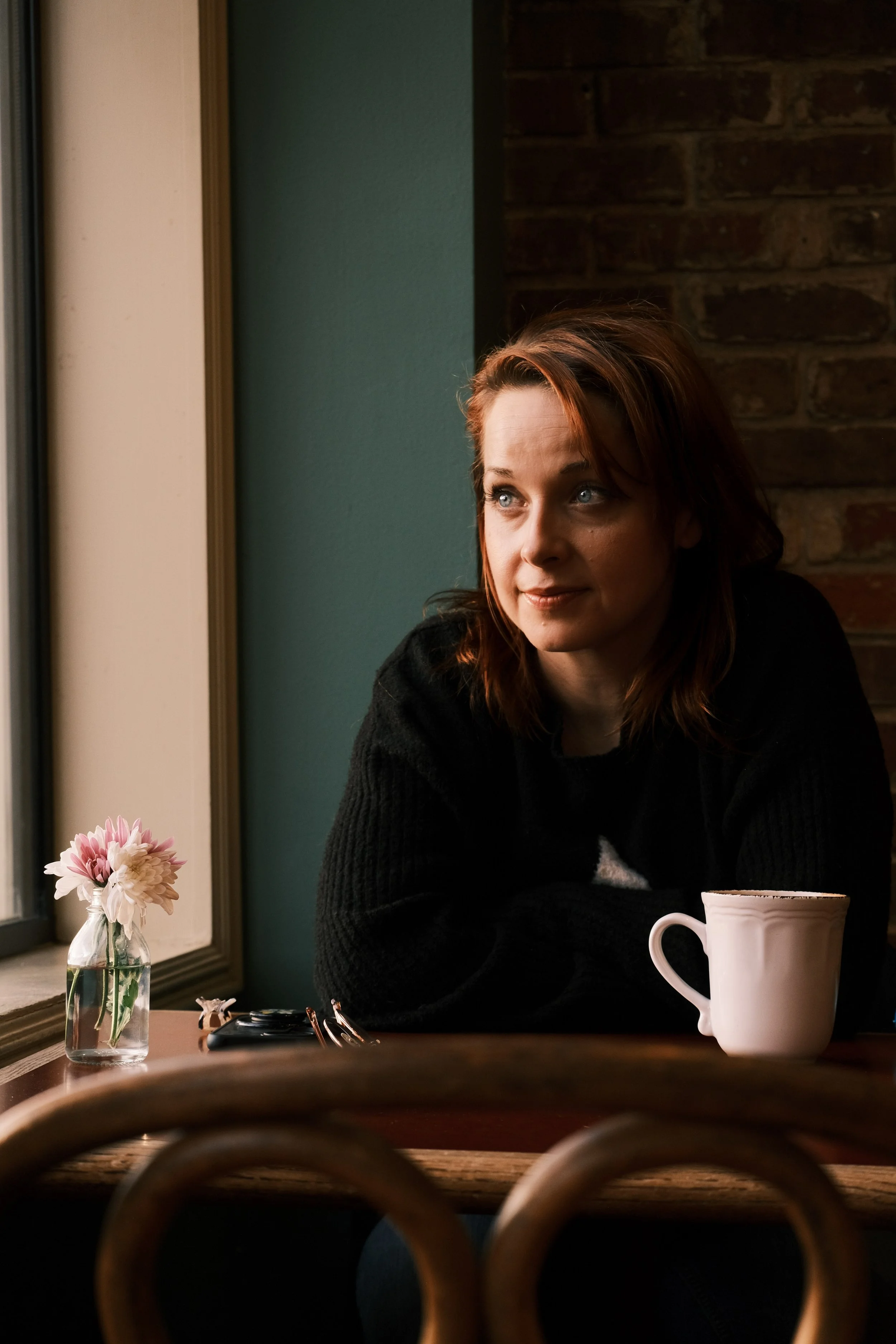 A woman with reddish-brown hair and blue eyes sitting by a window in a cafe, wearing a black sweater, with a white mug and a small vase with pink flowers on the table.