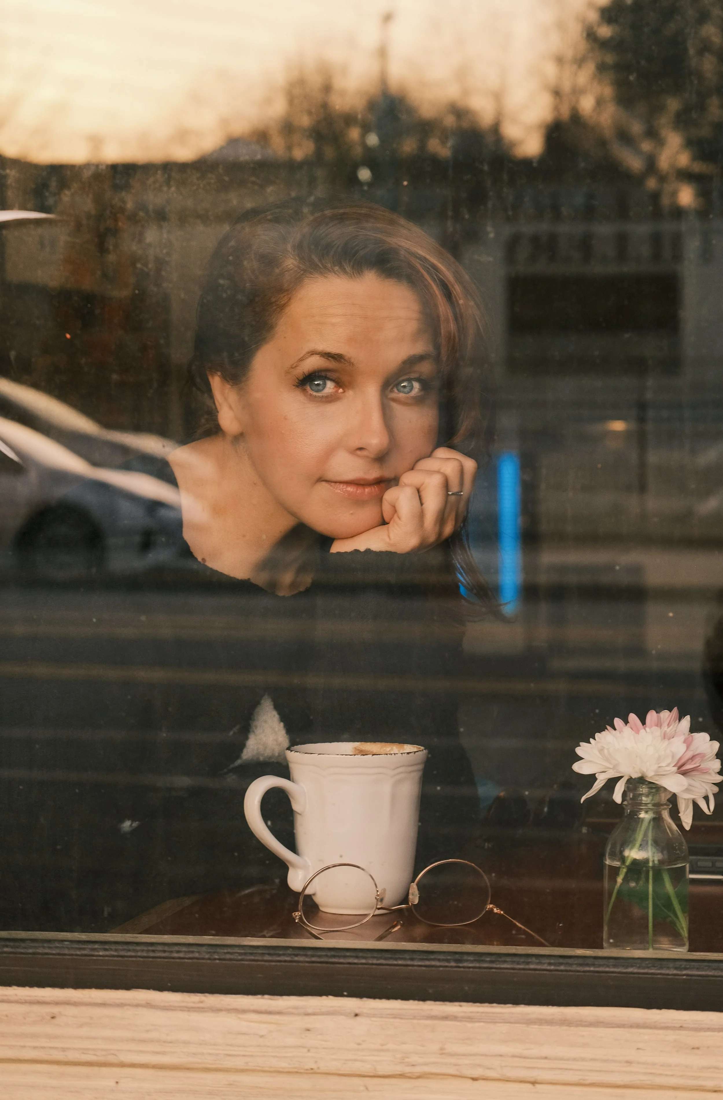 Woman looking through a window with a coffee mug, glasses, and a flower in a small vase on the table.