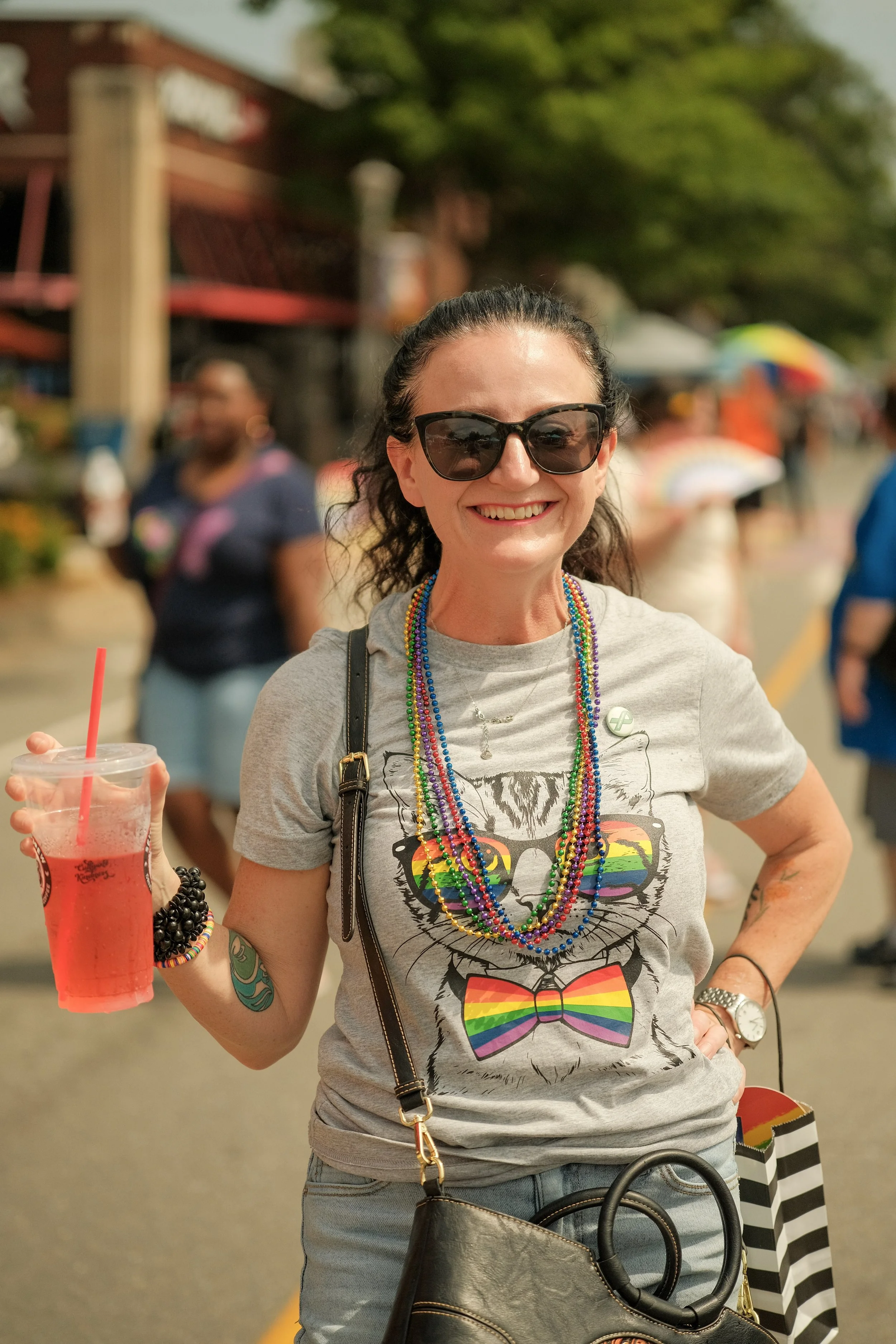 Woman smiling wearing sunglasses and rainbow-colored bead necklaces at a pride event, holding a red drink, with other people and colorful umbrellas in the background.