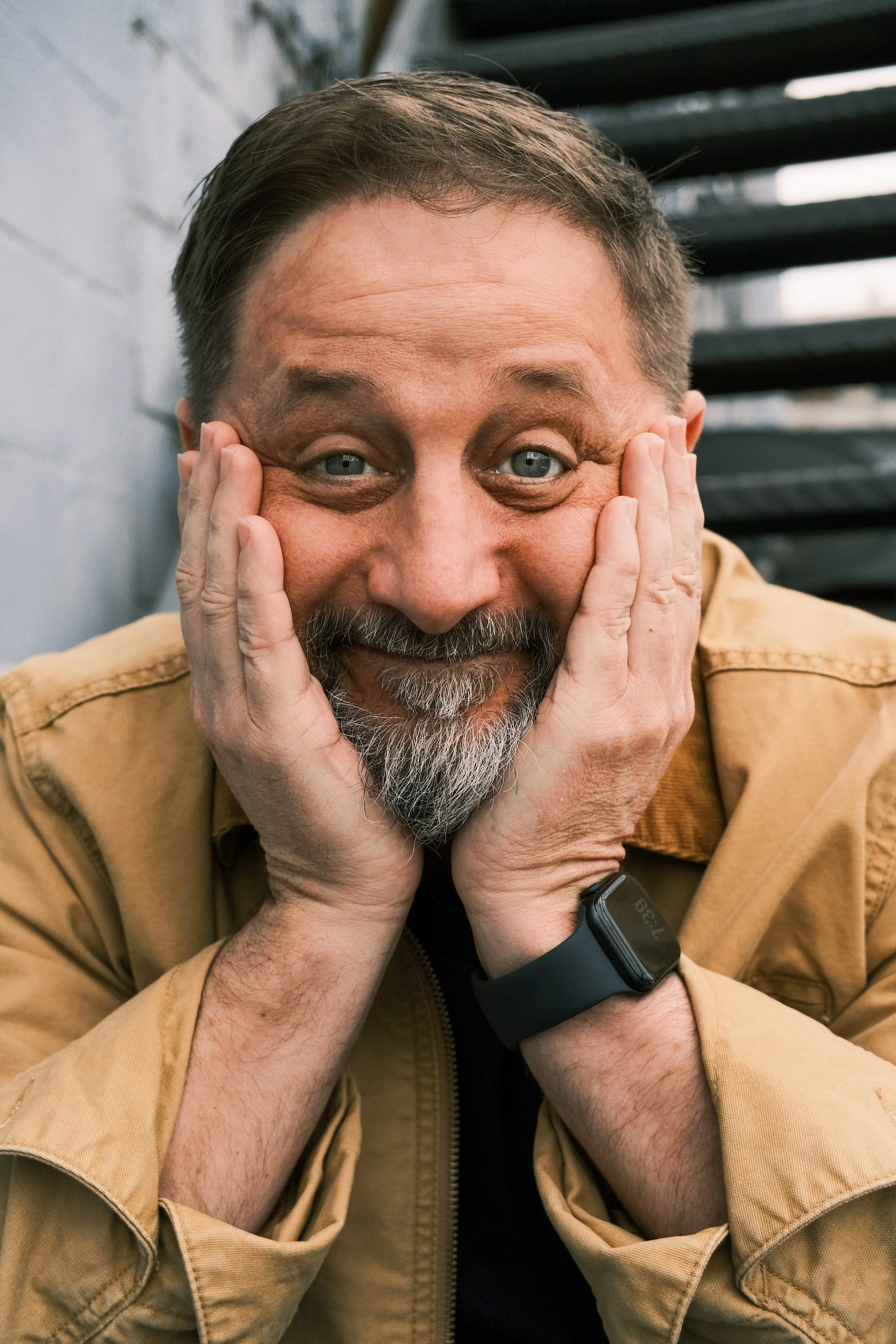 A man with gray hair and a beard is smiling with his hands on his face, eyes wide open, wearing a black wristwatch, and a tan jacket, against a gray wall and black metal steps.