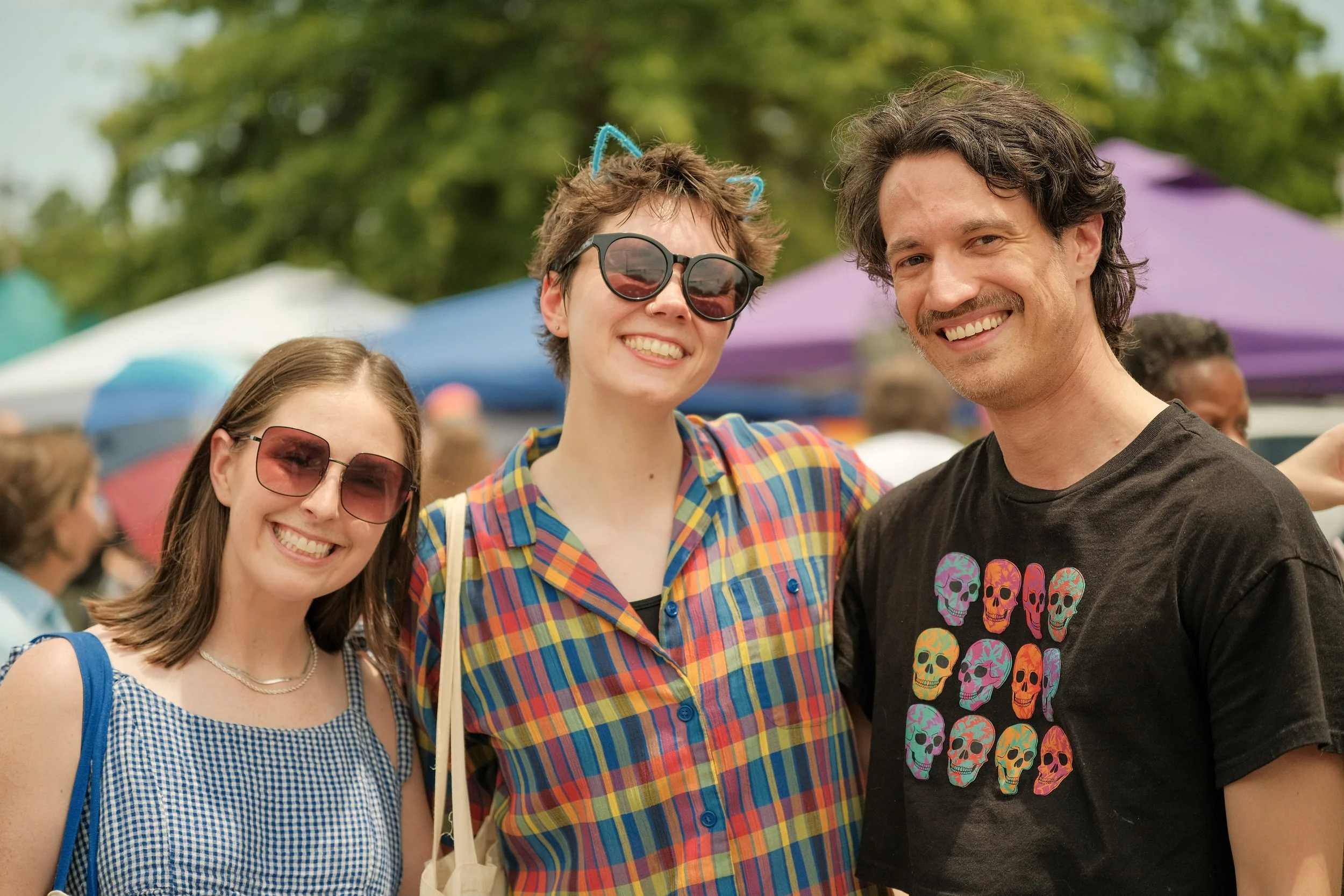 Three young adults smiling at an outdoor event with colorful umbrellas in the background, dressed casually.