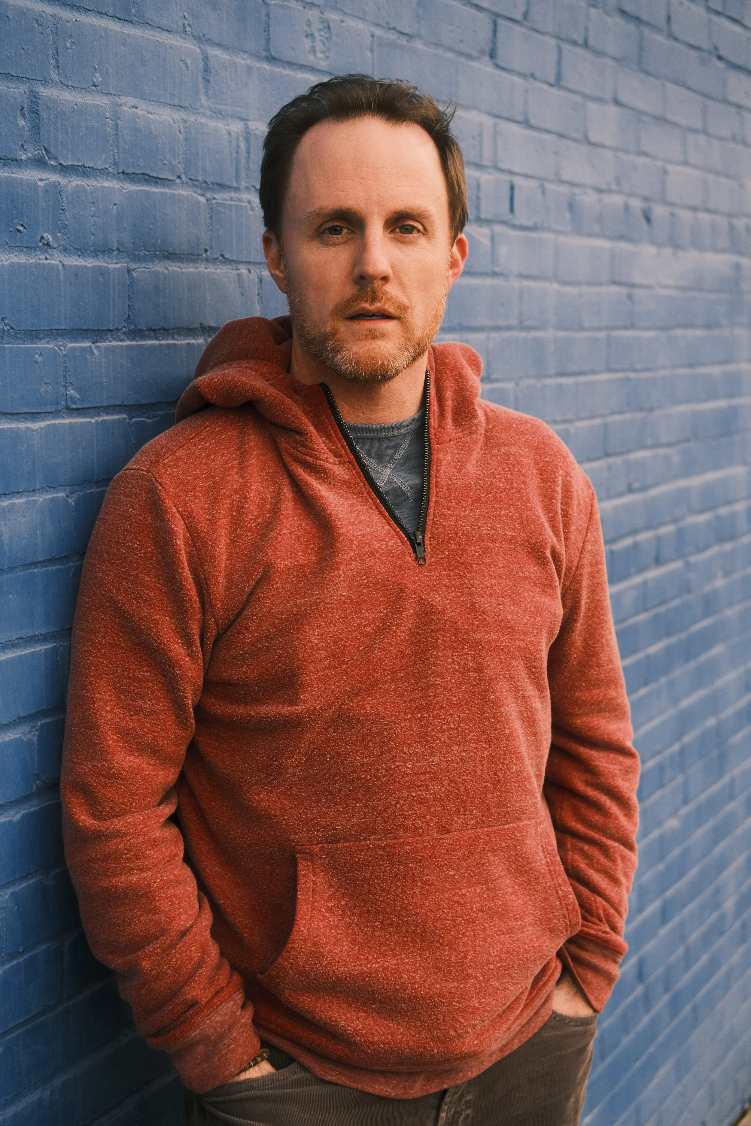 A man with brown hair and light facial hair leaning against a blue brick wall, wearing a red hoodie with a zipper and a dark shirt underneath.