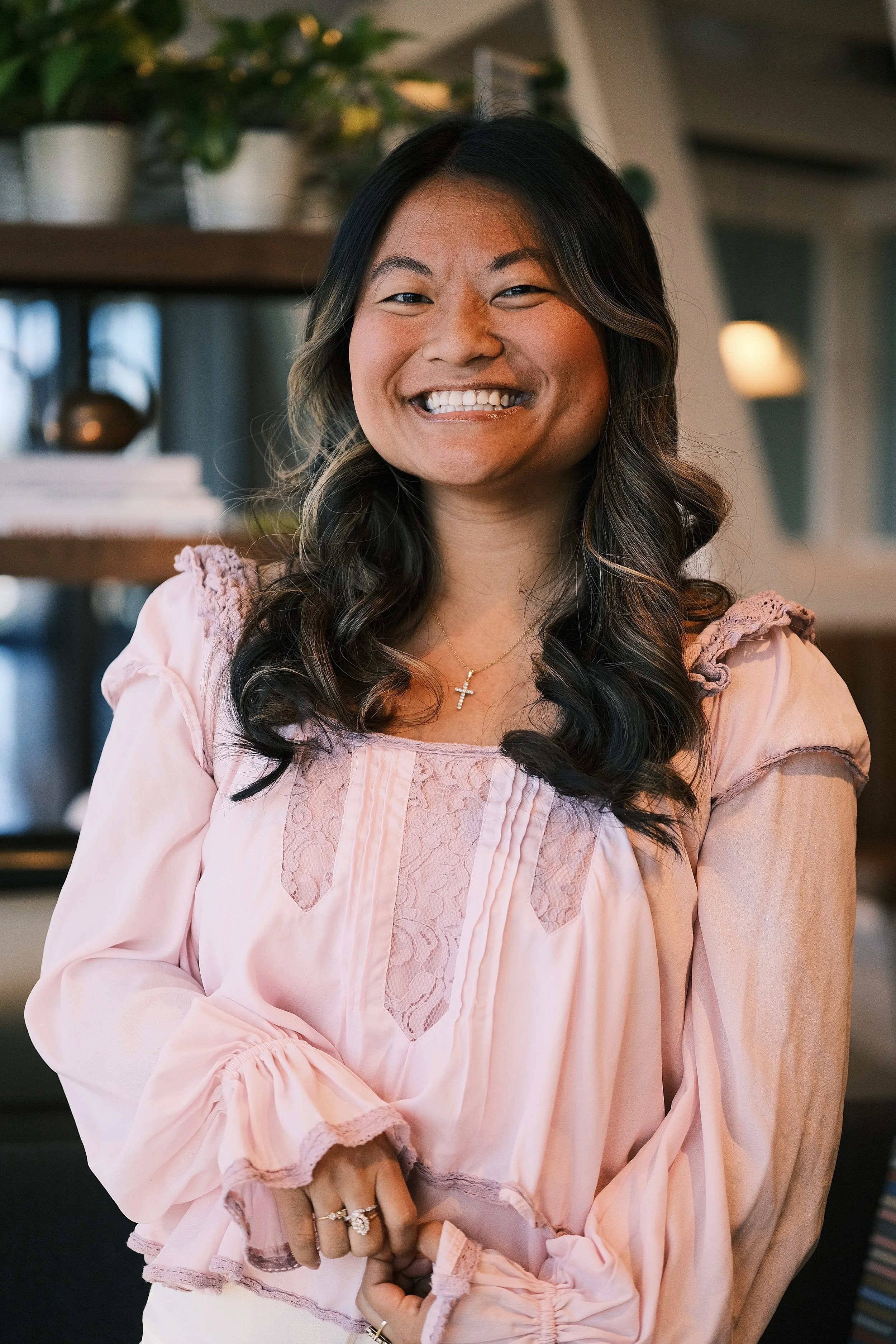 A woman with long, dark hair and glasses, smiling, wearing a pink blouse with lace details, indoors with shelves and plants in the background.