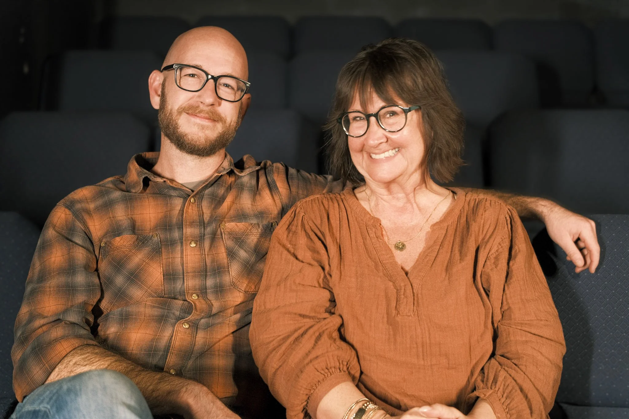 A man and woman sitting closely together on a dark theater seat, smiling. The man has a beard and glasses, wearing a brown plaid shirt. The woman has short, dark hair and glasses, wearing an orange blouse with jewelry. Their arms are around each othe