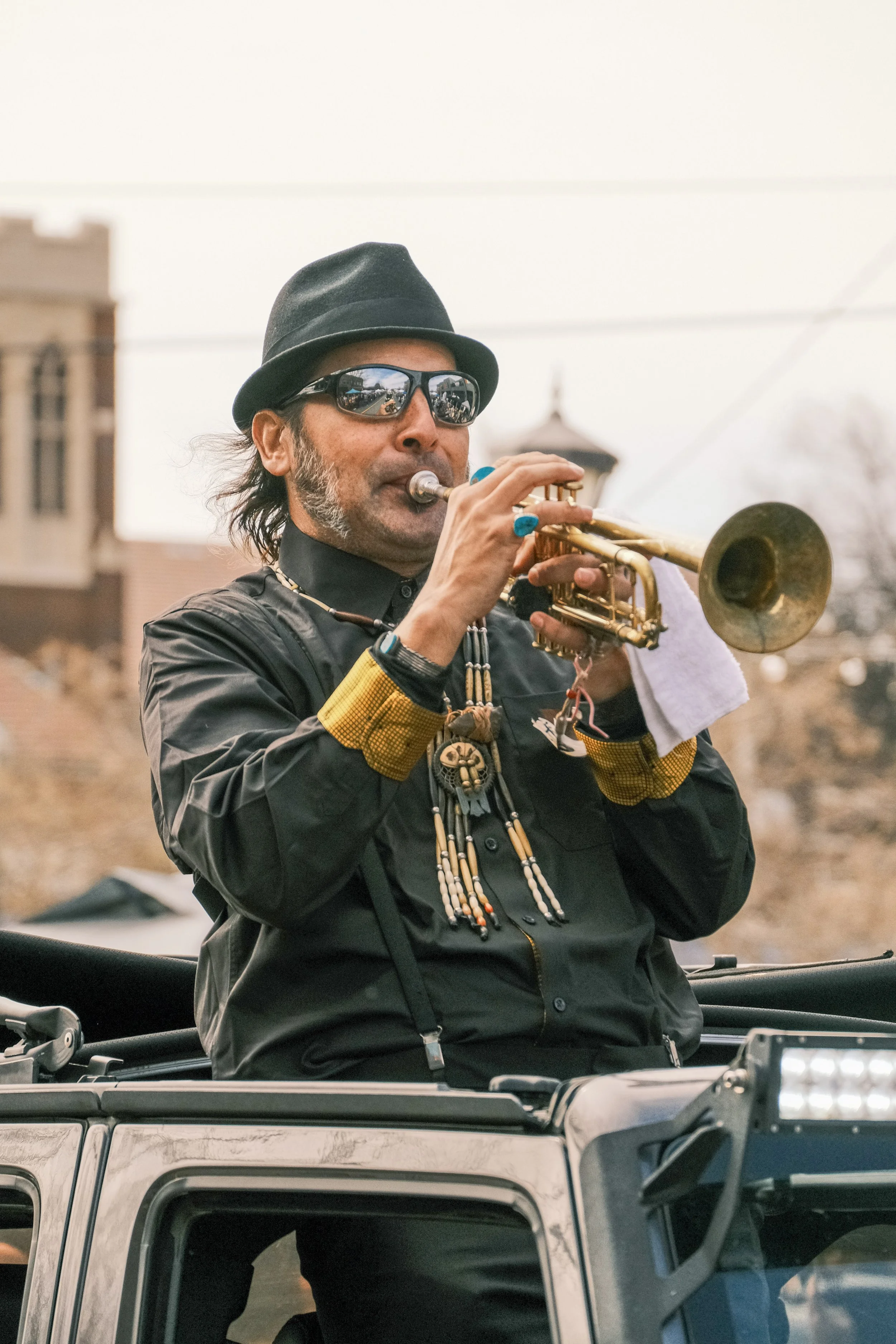 A man with dark hair and beard wearing sunglasses, a black hat, and a black jacket playing a trumpet outdoors.