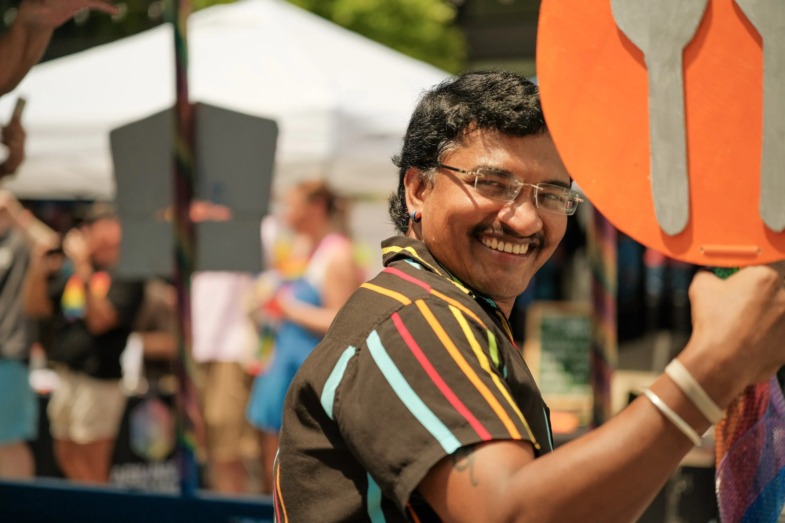 A man with curly black hair, glasses, and a colorful striped shirt smiling, surrounded by a crowd at an outdoor event with tents and colorful signs.