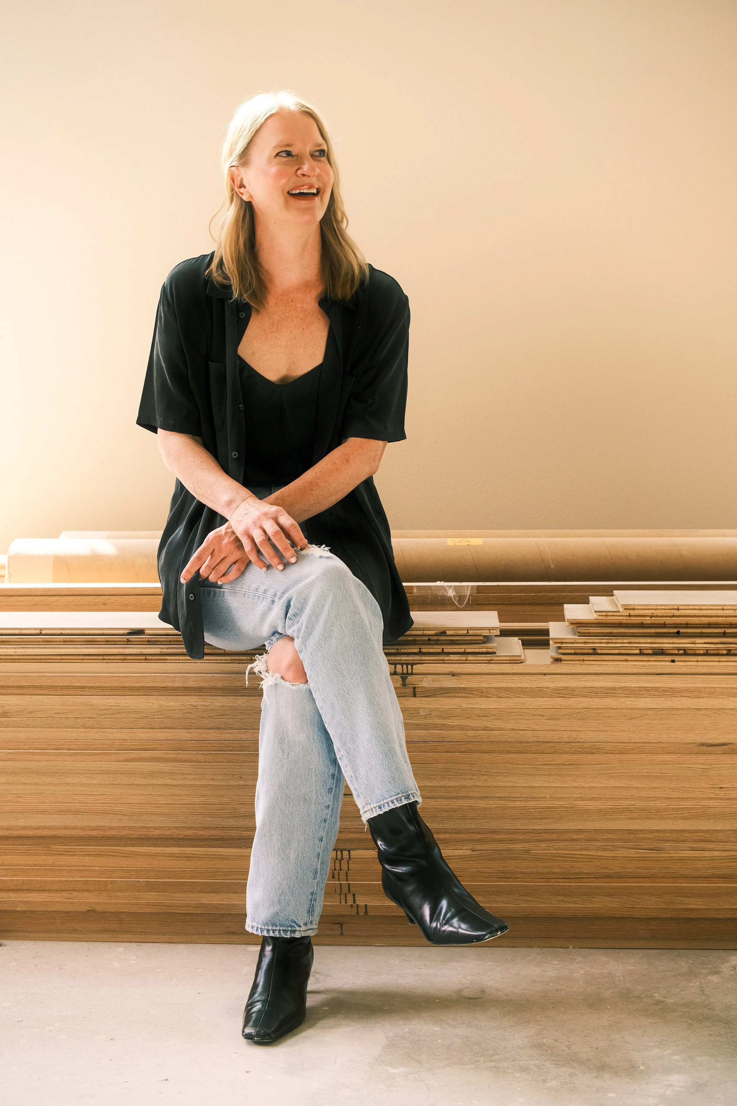 A woman with blonde hair sitting on a wooden bench, wearing a black top, gray ripped jeans, shiny black knee-high boots, and a black open shirt, smiling and looking to her right against a beige wall.