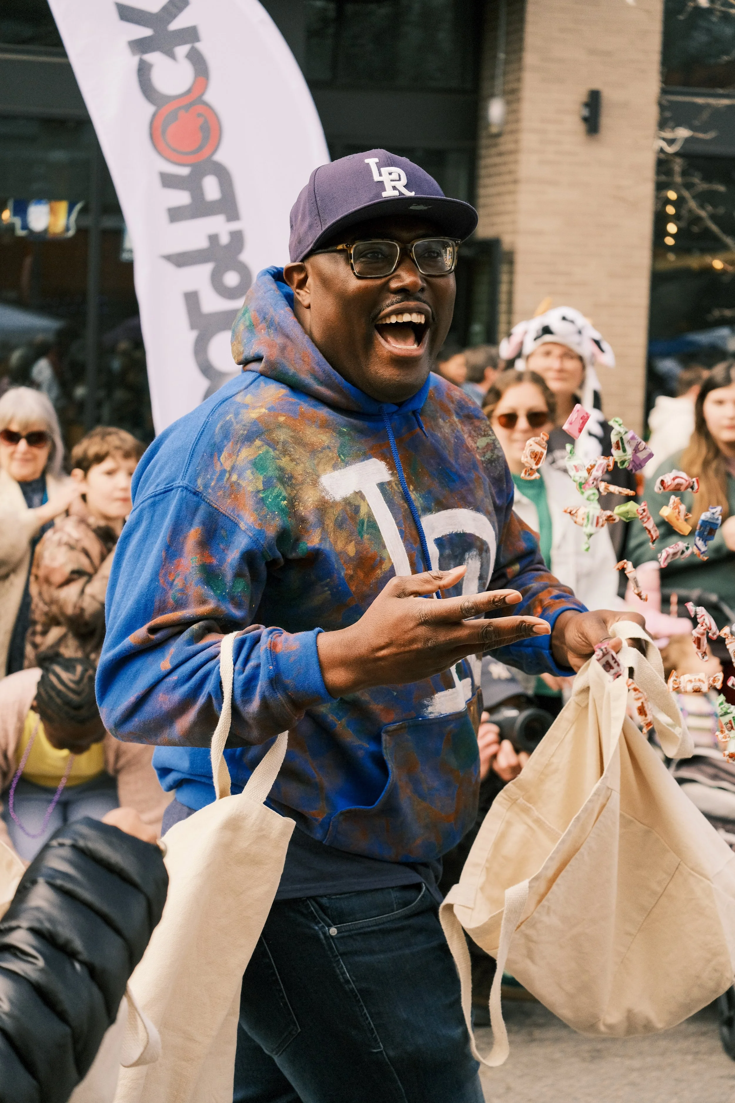 A man wearing glasses, a gray baseball cap with 'LR' on it, and a multicolored hoodie is smiling and holding a paper bag with candy around him at an outdoor event, with a crowd of people and a vertical sign in the background.