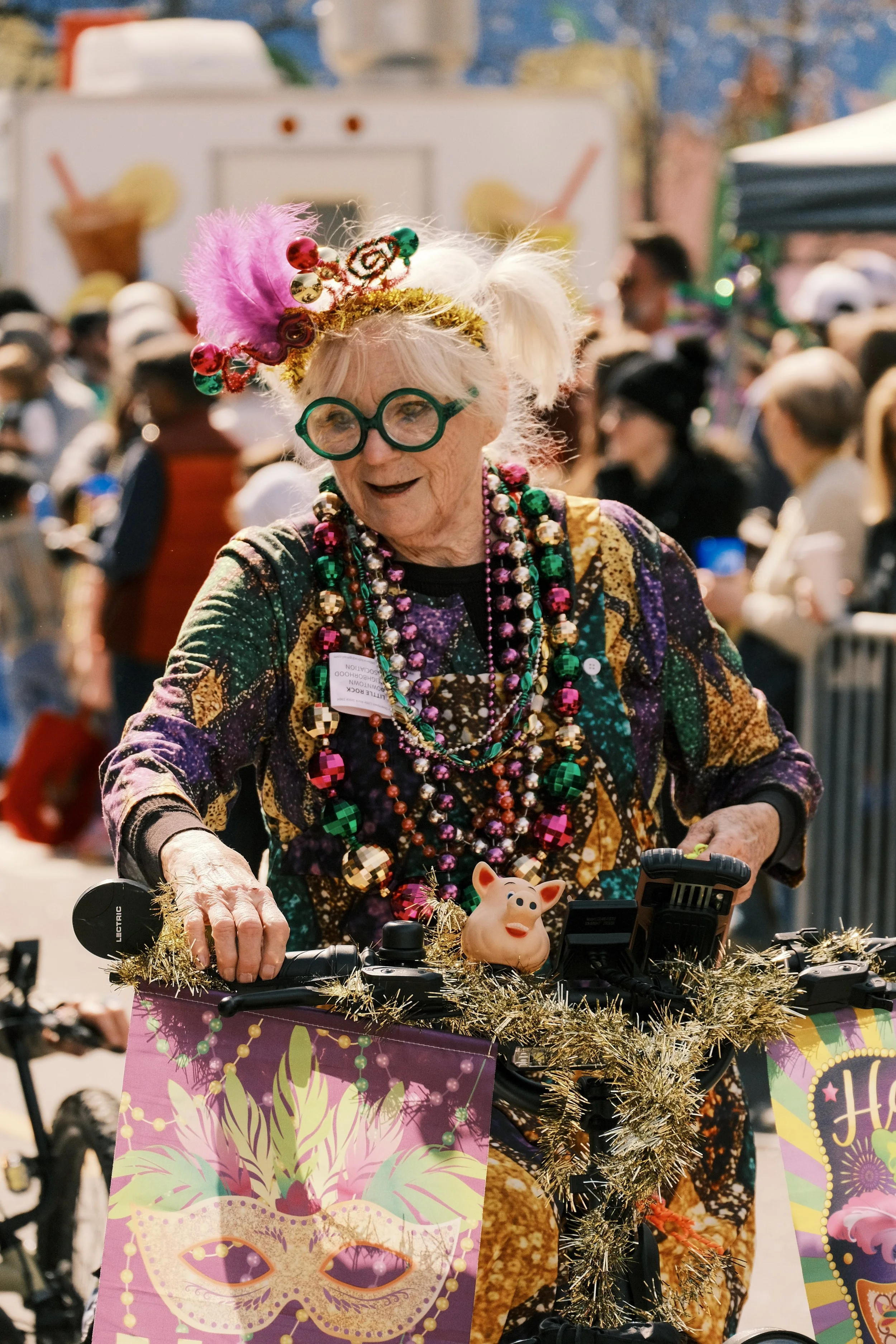 An elderly woman dressed festively with colorful beads, a decorative headband, and round green glasses, riding a decorated bicycle at a Mardi Gras or carnival celebration with a crowd and festive decorations in the background.