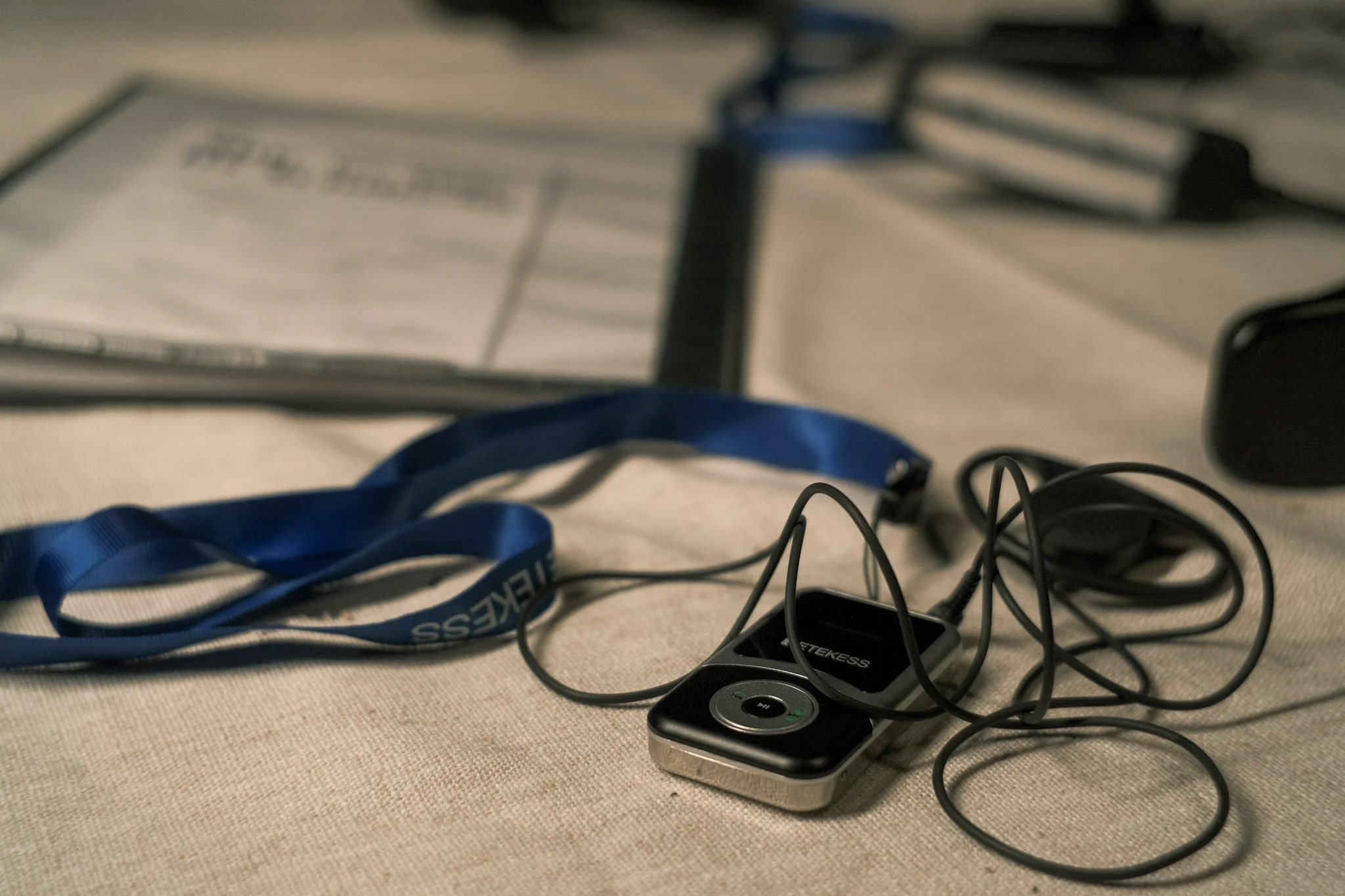 Unorganized electronics and accessories on a beige surface, including a black wireless speaker, tangled black and gray cables, a blue lanyard, and a plastic item with a small screen.