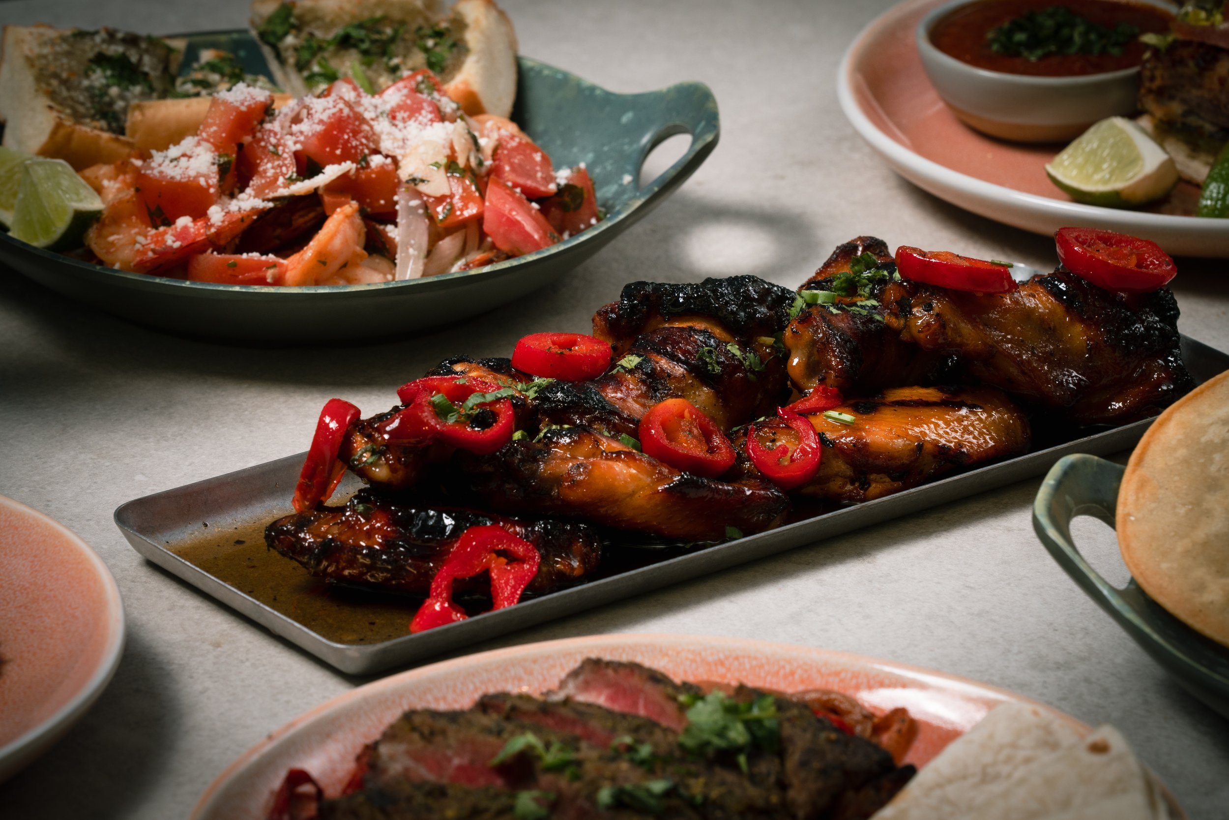 Mexican dishes including grilled chicken with red chili slices and chopped herbs, chopped tomato and onion salad with grated cheese, and traditional Mexican sauces with lime wedges.