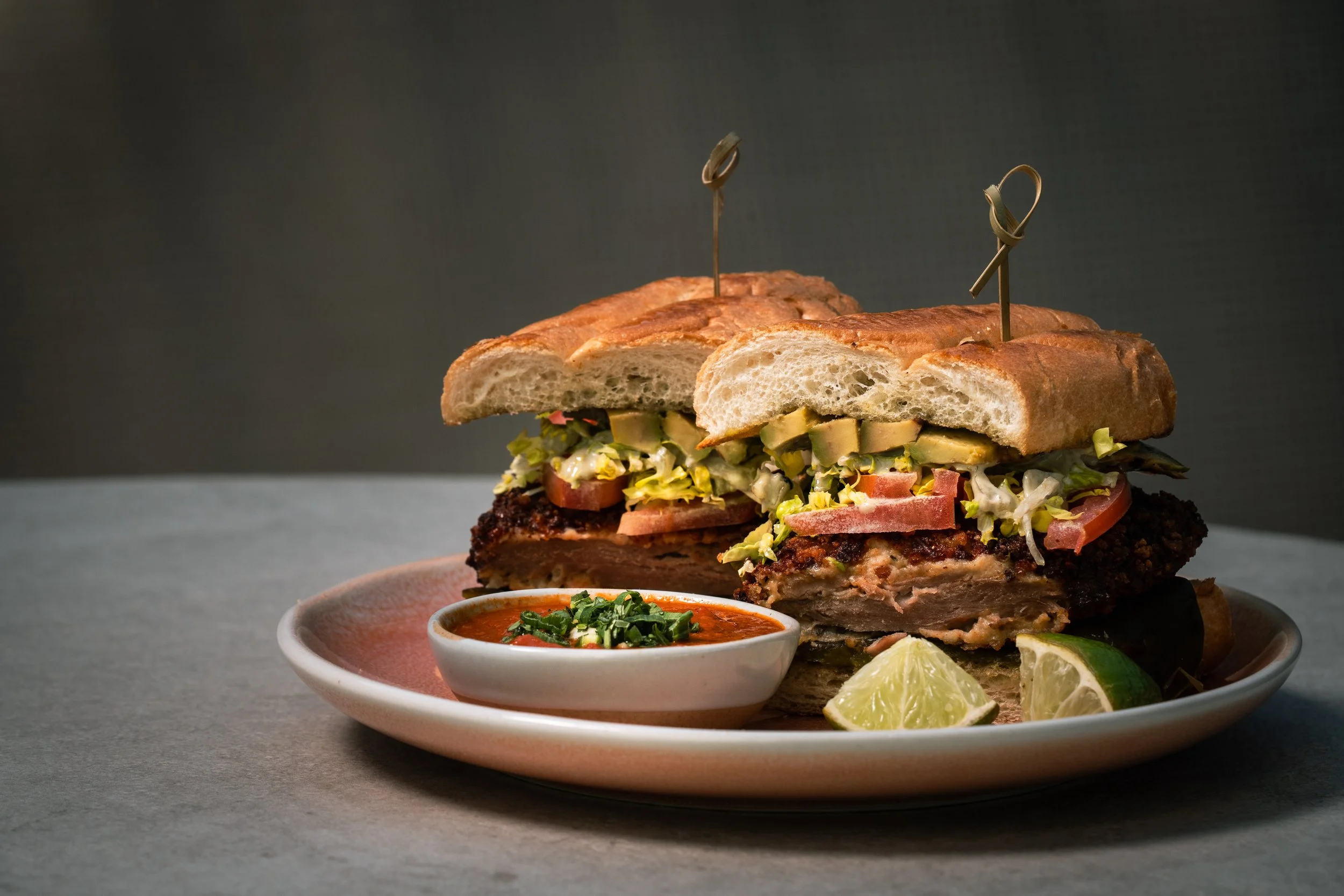 A sliced sandwich with various ingredients on a plate, served with lime wedges and a small bowl of dipping sauce.
