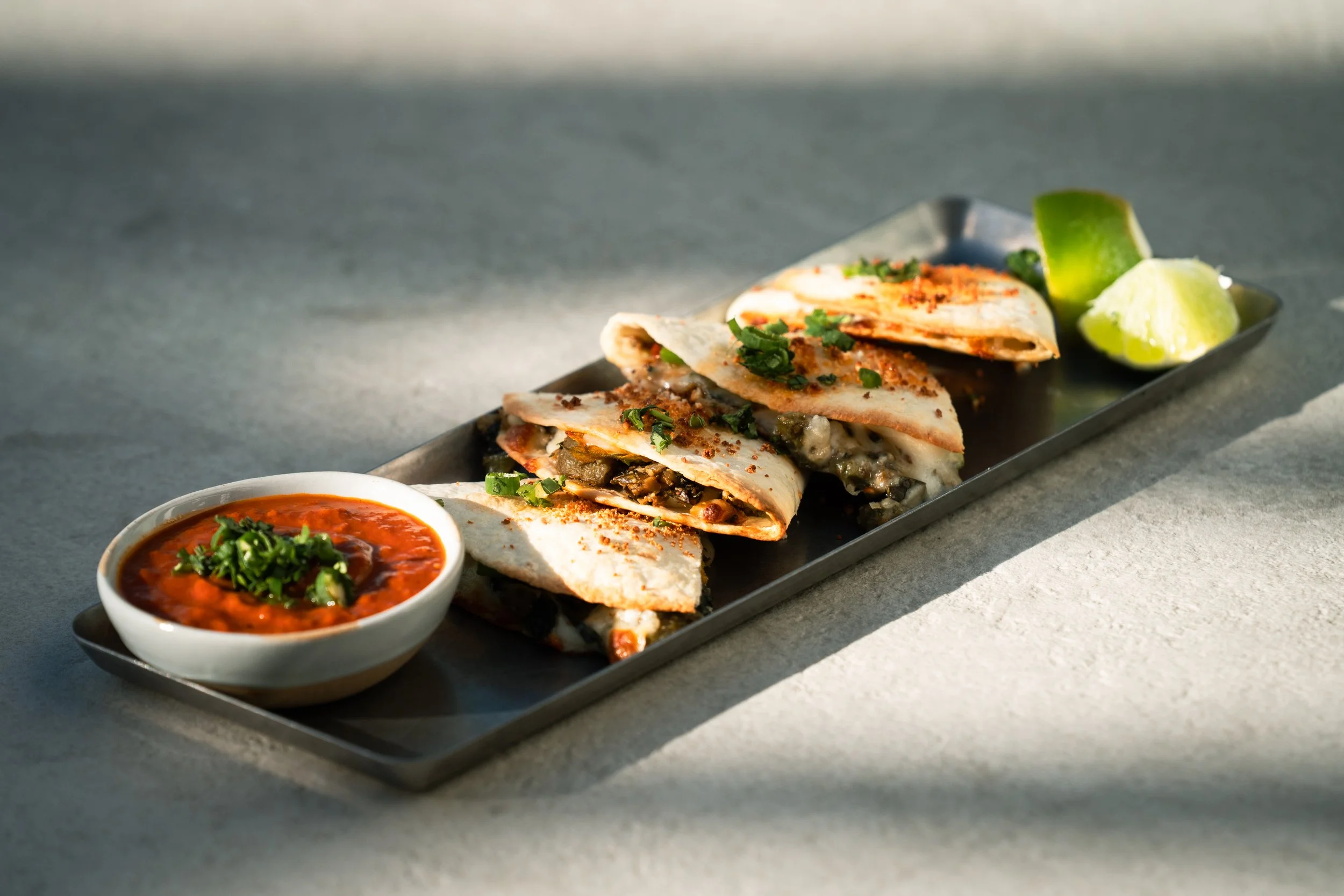 Close-up of a black rectangular plate with four slices of flour tortilla, queso chihuahua, nopales, lacinato kale, garlic, cotija, salsa morita.