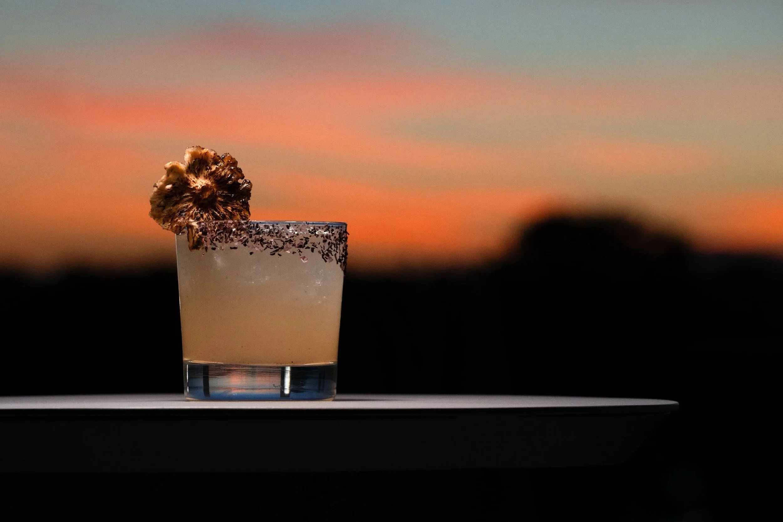 A cocktail with a frosted rim topped with a decorative flower garnish against a colorful sunset sky.