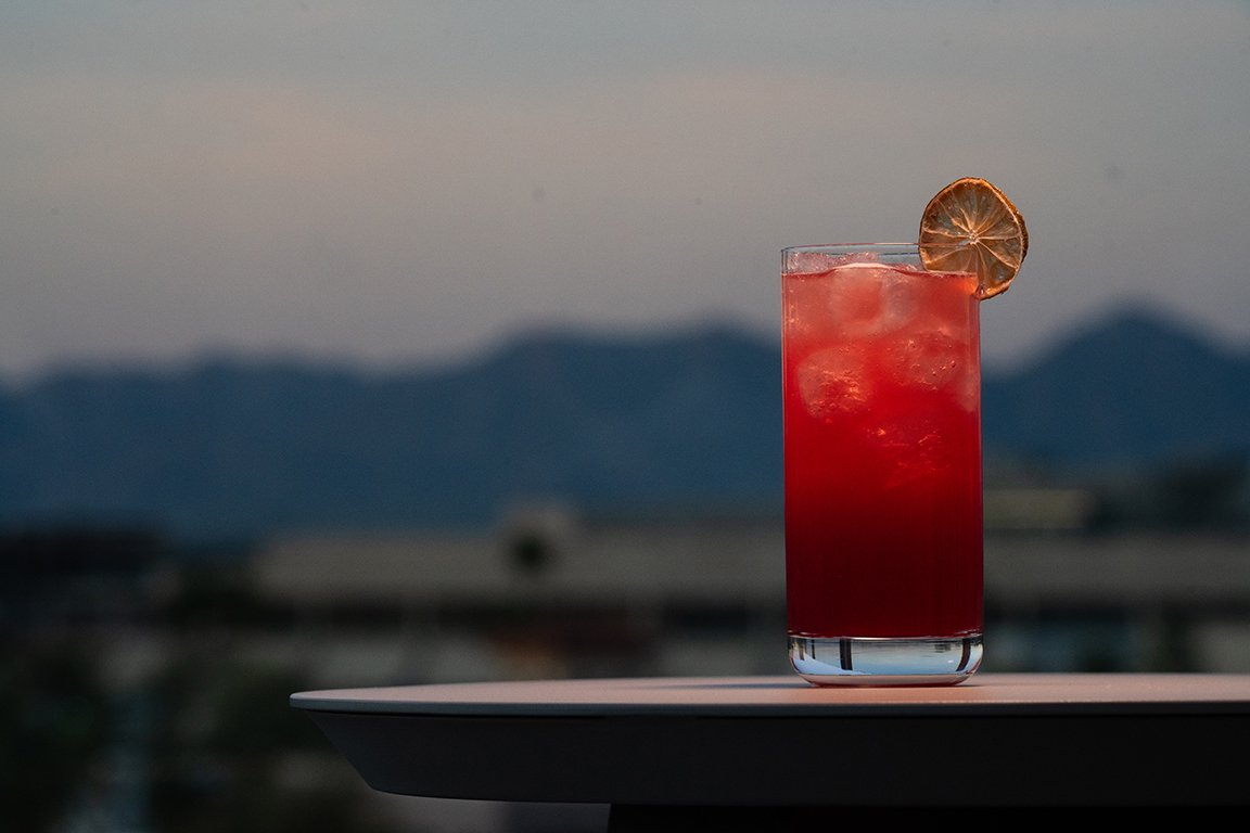 A tall glass filled with a red beverage and ice cubes, garnished with a dried lemon slice on the rim, placed on a white surface outdoors with mountains in the background during evening or dusk.
