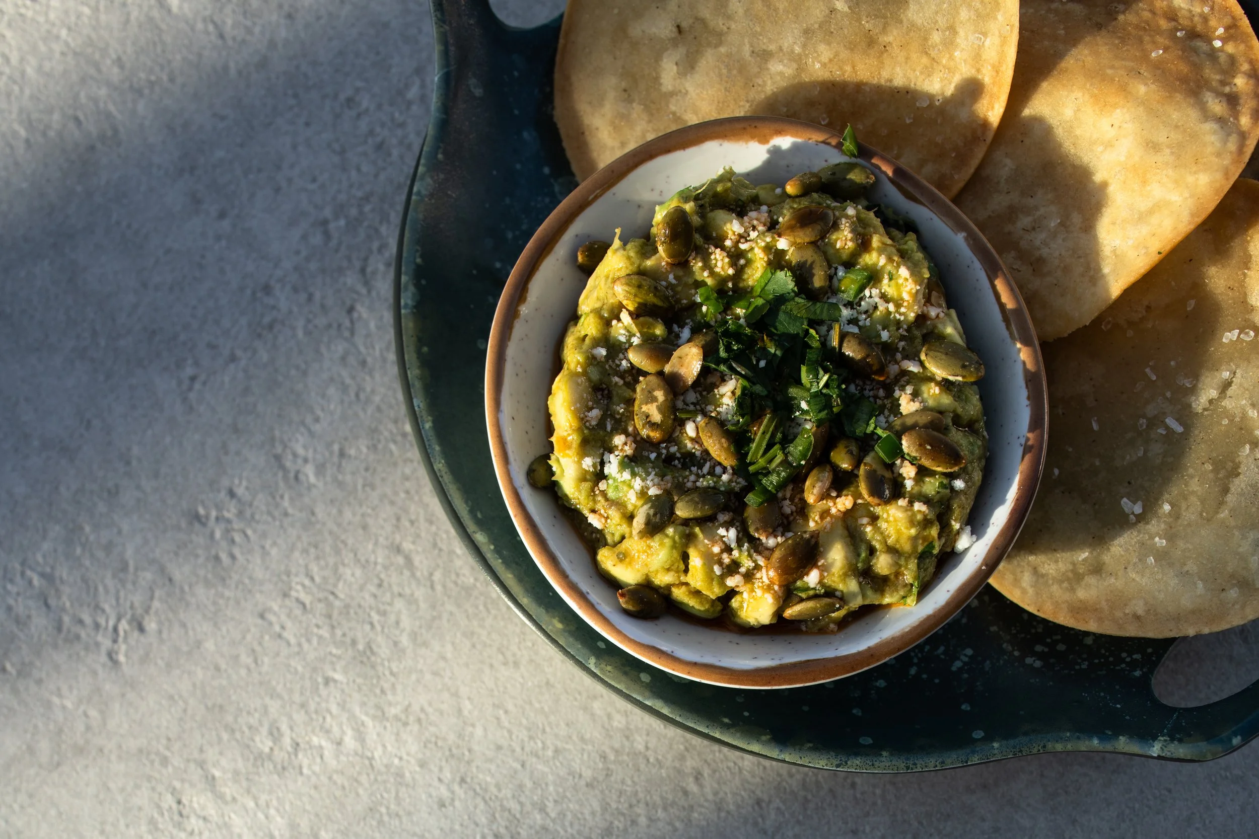 Small bowl of squashed avocado topped with cilantro, garlic serrano, green onion, cotija, salsa seca, placed on a plate with mini tostadas.