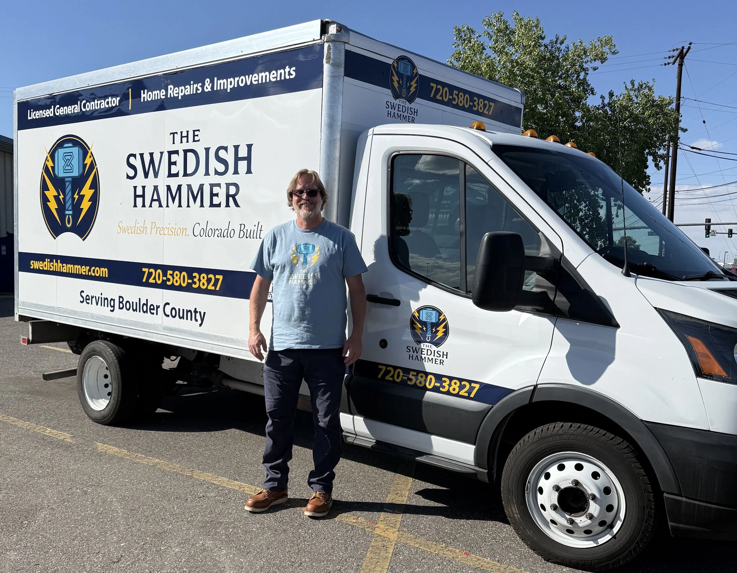 A man standing in front of a white service van with the logo and branding for The Swedish Hammer, a home repair and improvement company. The man is wearing sunglasses, a gray T-shirt with the company's logo, dark pants, and brown shoes. The van displays the company's contact information and website on the side, and the background shows a partly cloudy sky, green trees, and utility poles.