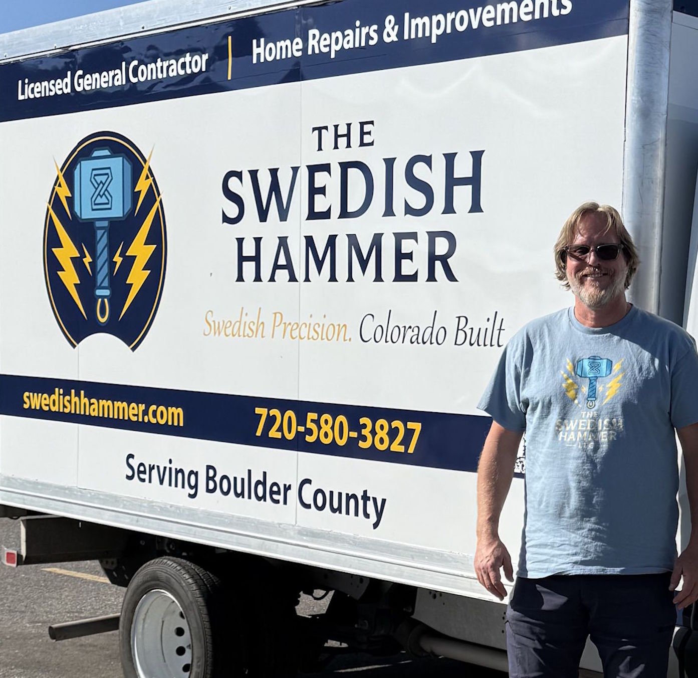 A man wearing sunglasses and a gray T-shirt with a logo, standing next to a mobile advertisement trailer for The Swedish Hammer, a home repair and improvement business. The trailer displays the company’s name, logo, slogan, website, and phone number. The background shows a clear sky.