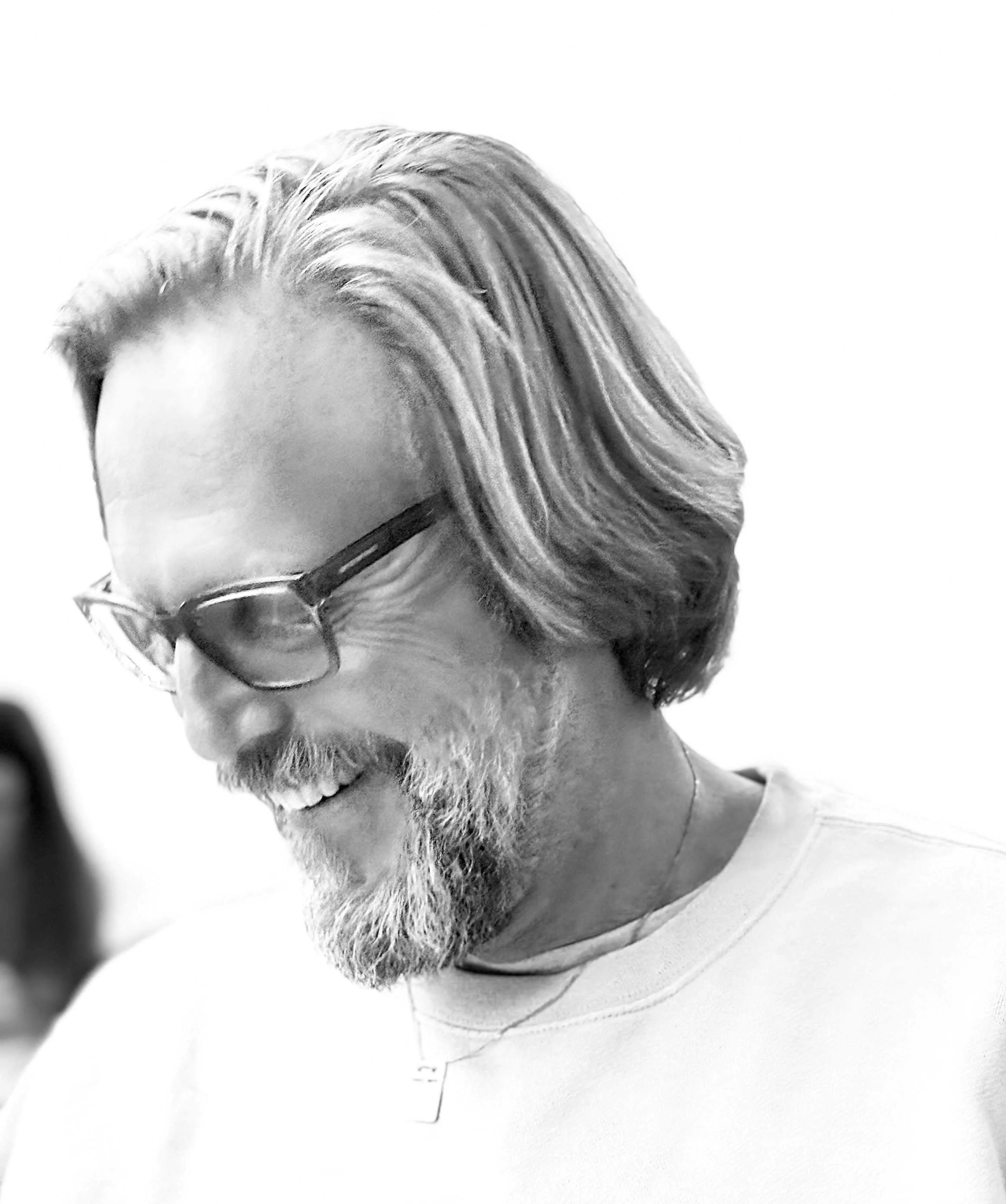 A man with long, wavy hair and a beard is smiling, wearing glasses, and a light-colored shirt.