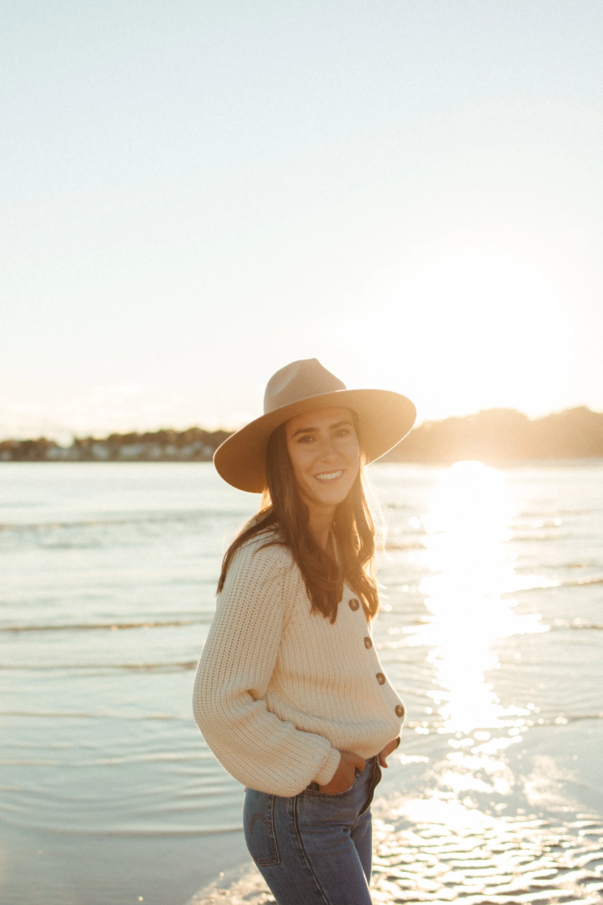 A woman standing on the beach during sunset, wearing a wide-brimmed hat and a cream-colored sweater, smiling at the camera.