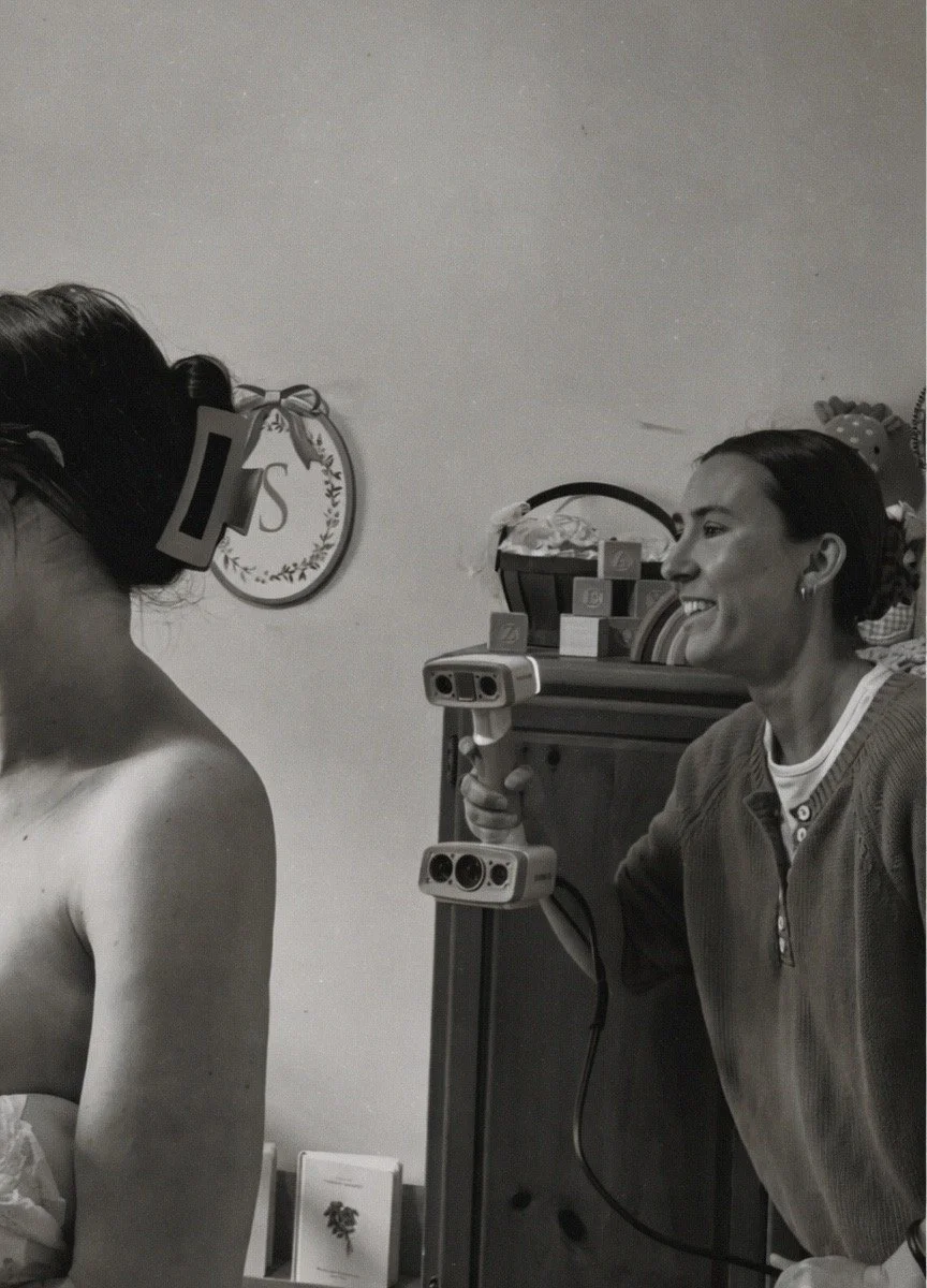 A woman holding a video camera on her shoulder, smiling as she films another woman with a wedding veil and a strapless dress, in a room decorated with children's items and framed artwork.