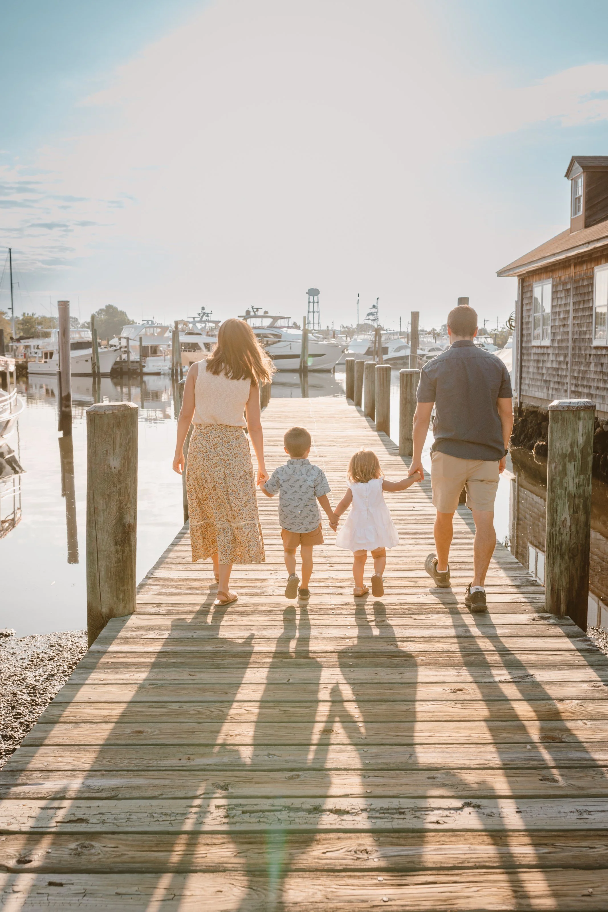 Family of four walking on a wooden dock towards boats at a marina during sunset.