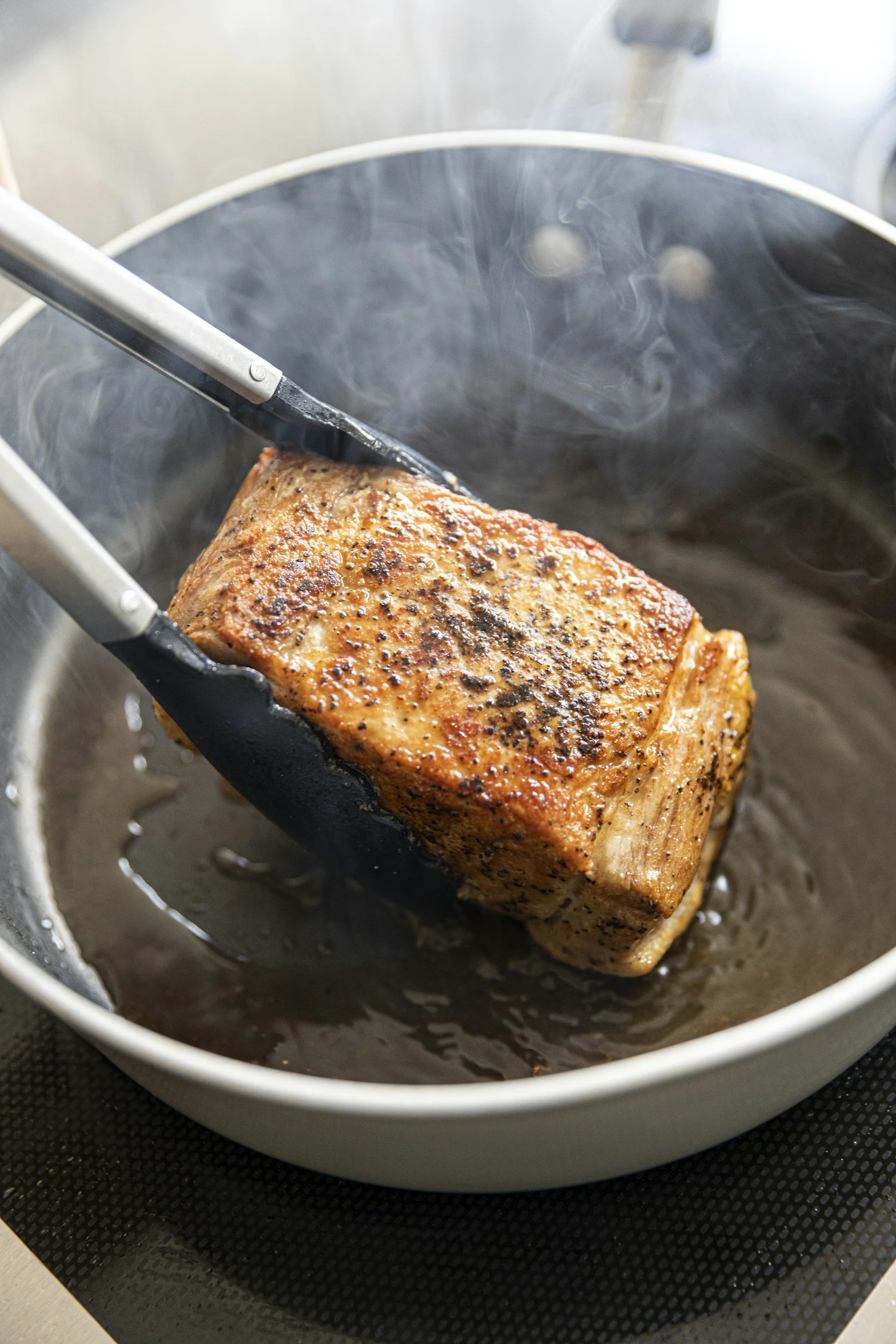 A piece of seasoned, cooked meat frying in a pan with smoke rising from it.