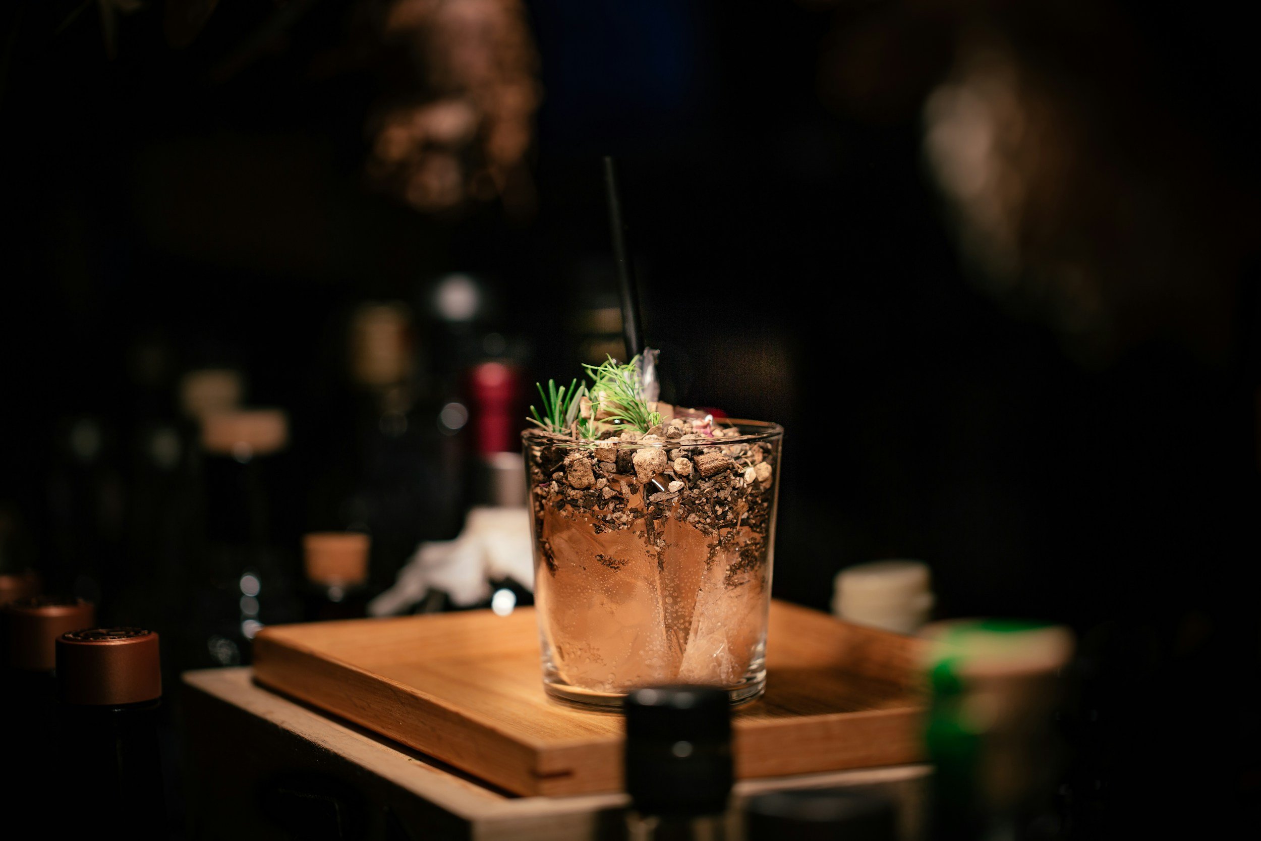 A cocktail drink in a glass with herbs and toppings, on a wooden board in a dimly lit setting.