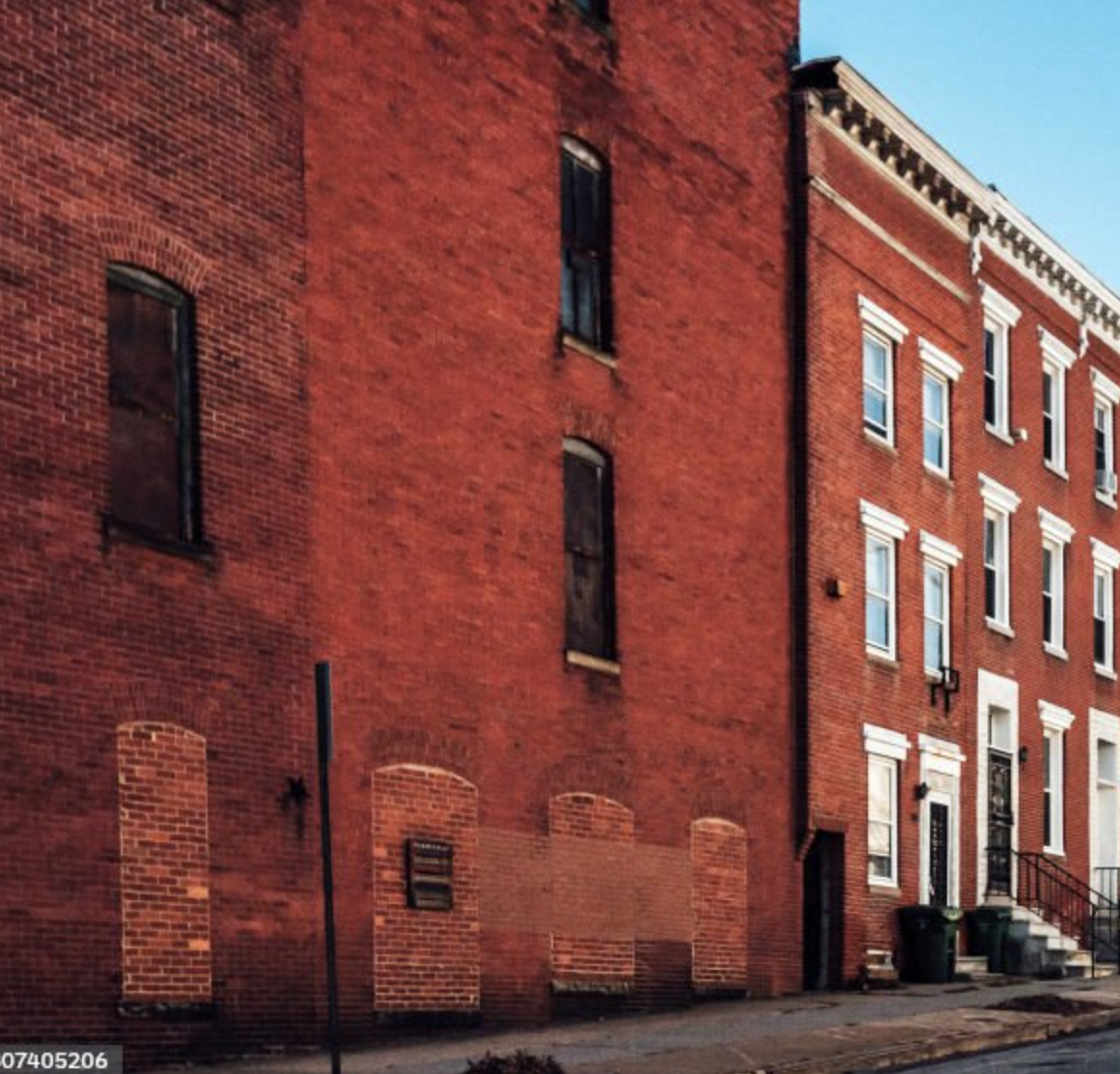 A street view of red brick row houses with multiple windows, steps leading to front doors, and trash bins outside. The buildings have decorative white trim and cornices under a clear blue sky.