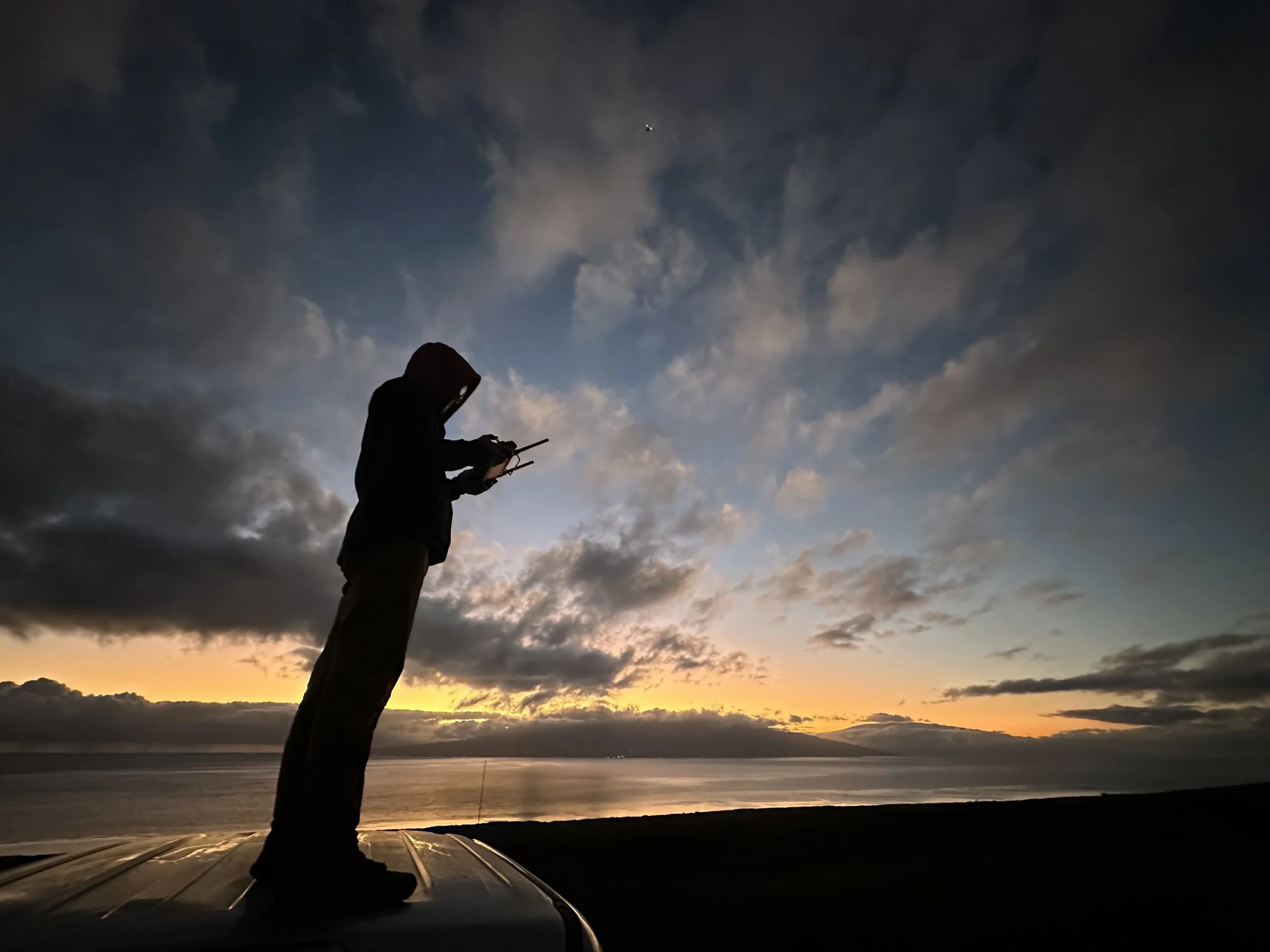 Silhouette of a person standing on top of a vehicle at sunset, holding a drone controller and looking at a drone in the sky, with clouds and a calm body of water in the background.