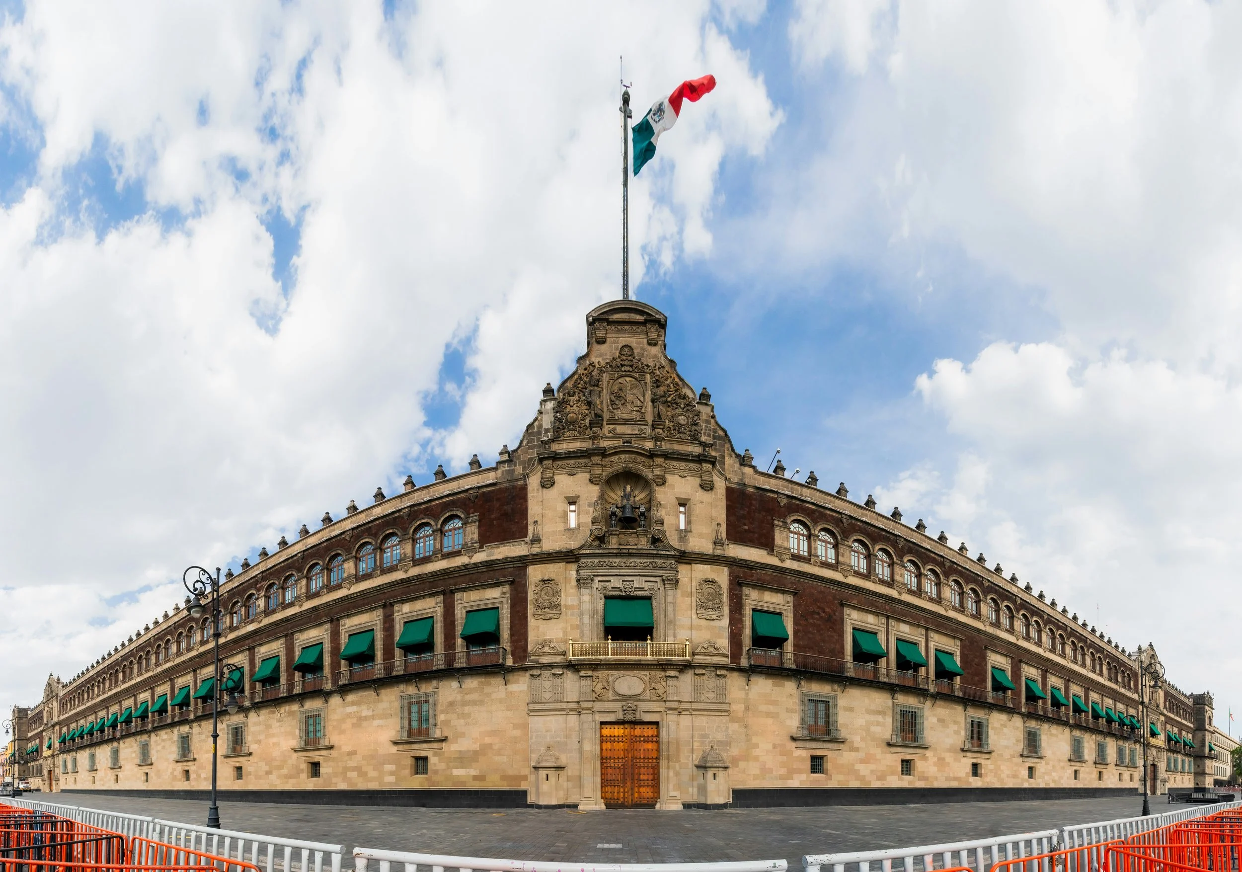 The image shows the National Palace in Mexico City, a large historic building with a corner tower, decorated with a Mexican flag on top, green window awnings, and detailed stonework. The sky is partly cloudy with some blue sky visible.