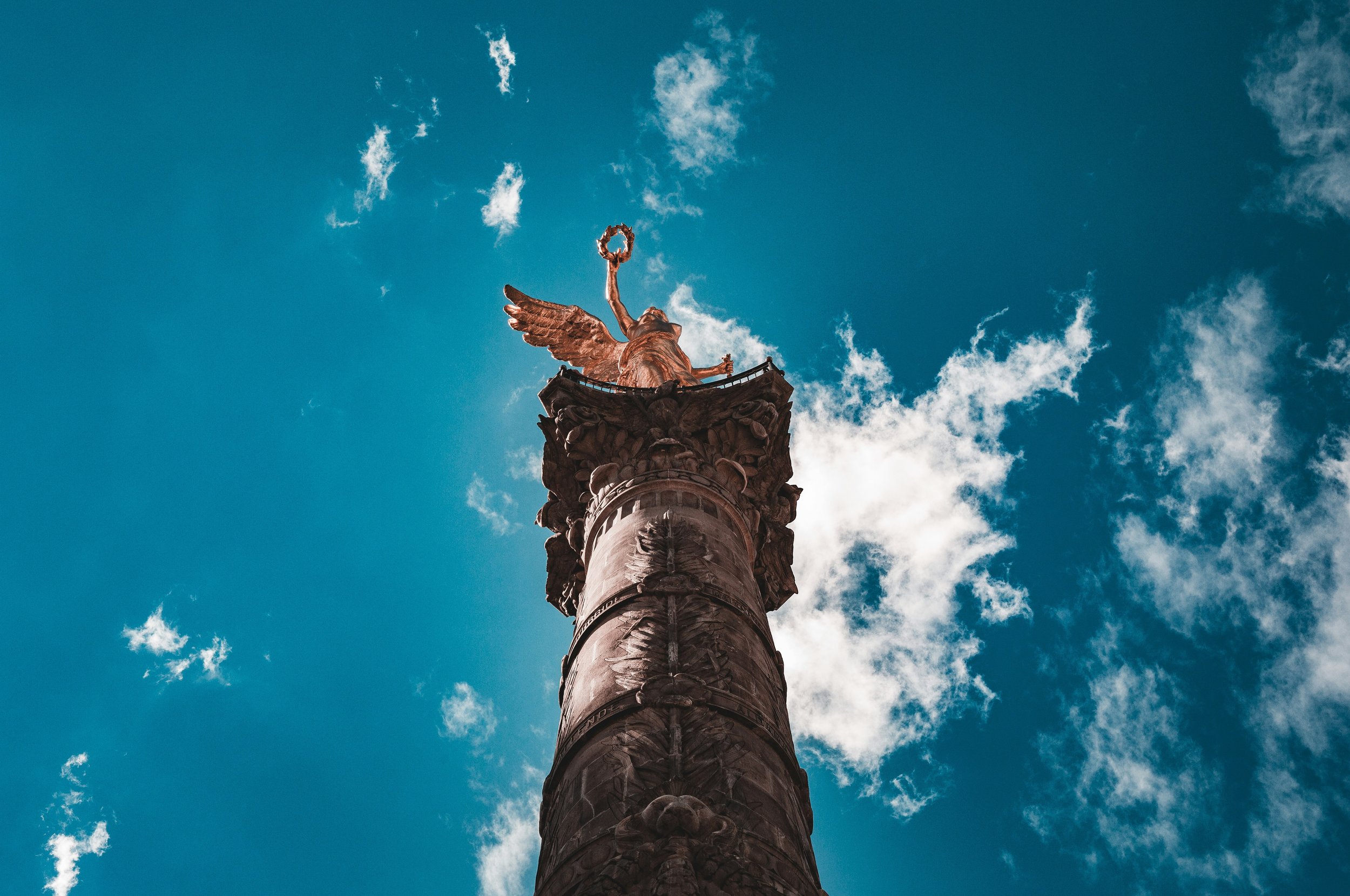 A tall monument topped with a golden angel holding a wreath, set against a blue sky with scattered clouds.