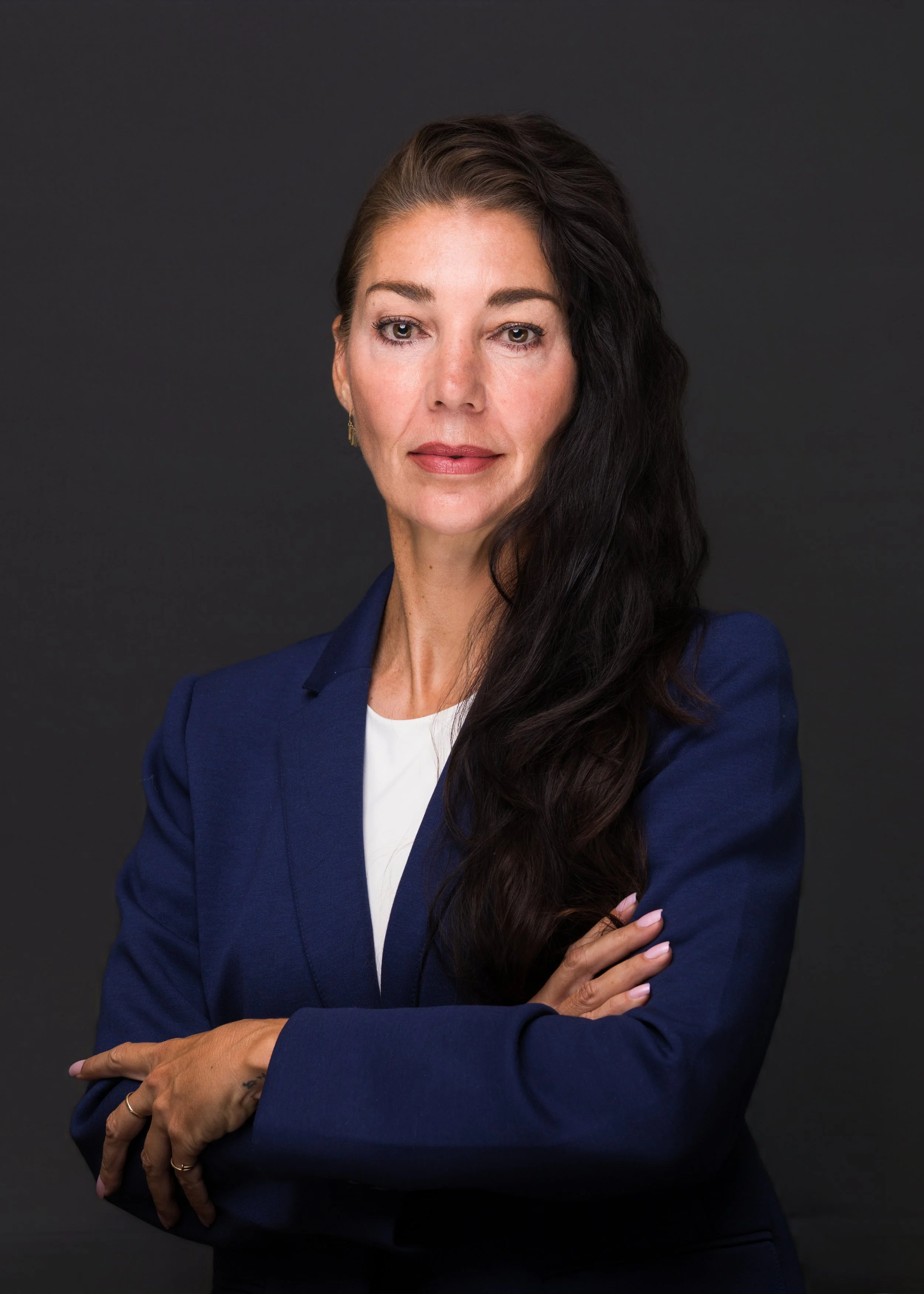 Portrait of a woman with long dark hair, wearing a navy blazer over a white top, standing with arms crossed against a dark background.