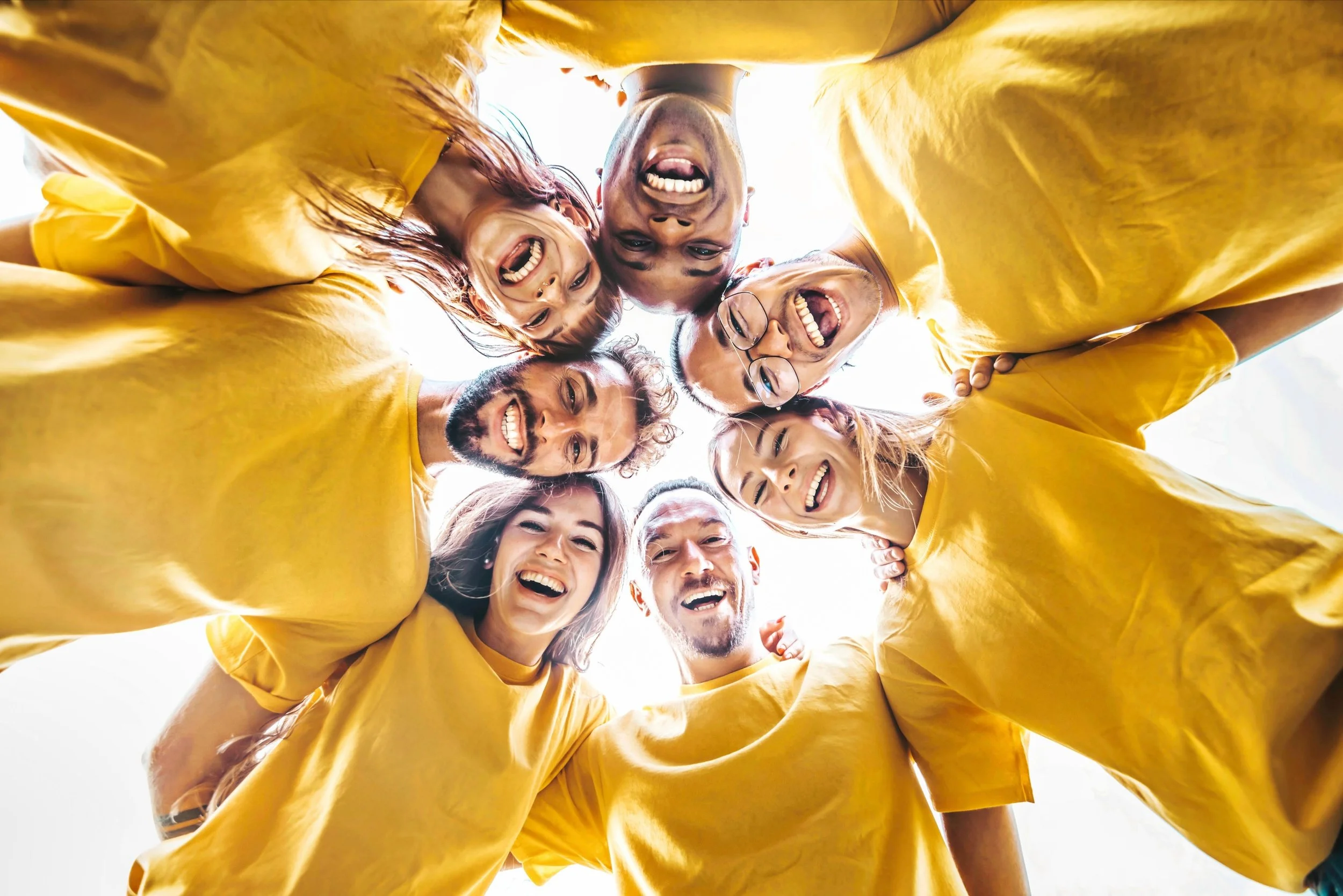 A diverse group of people wearing yellow shirts are standing in a circle, looking down at the camera and smiling widely.
