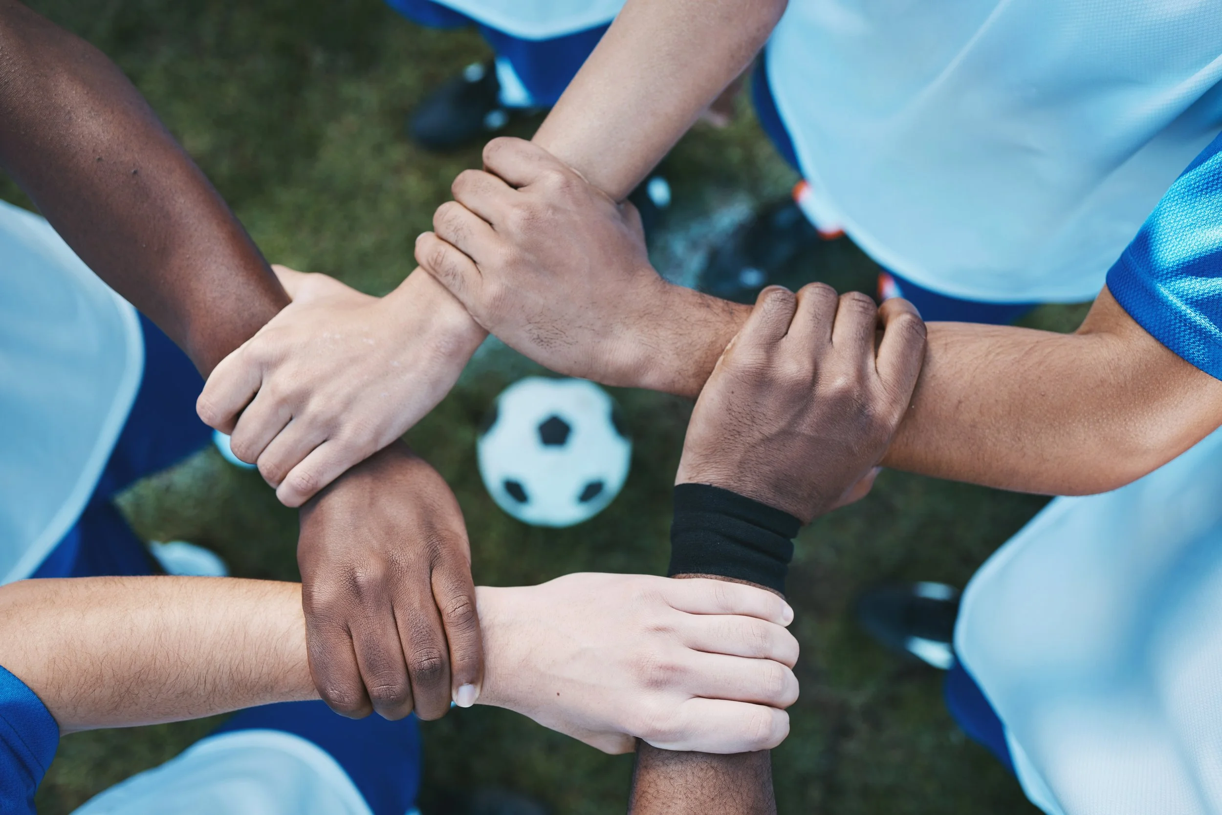 Group of diverse soccer players putting their hands together in a team huddle with a soccer ball on the grass below.