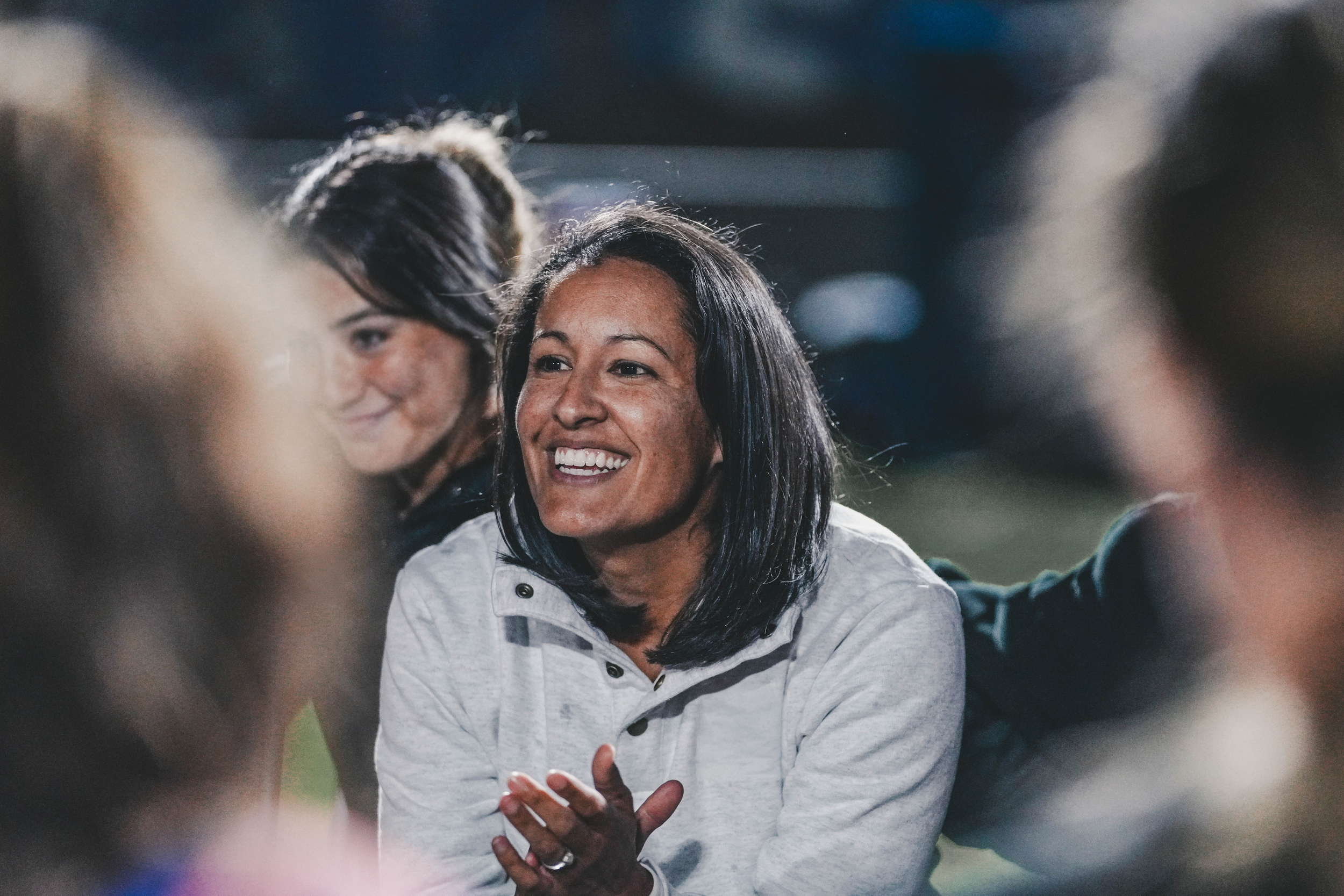 A woman with black hair smiling and clapping during an outdoor event at night, surrounded by other people.