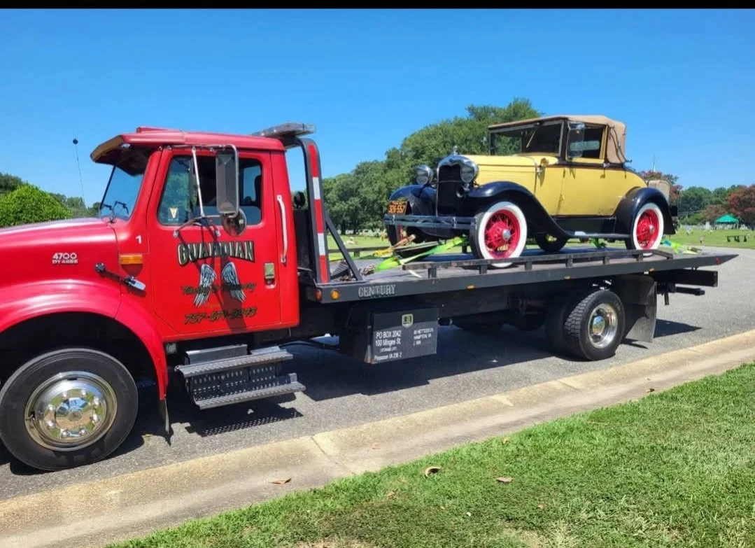 A red tow truck parked on the side of a road, carrying a vintage yellow and black car with red wheels on its flatbed.