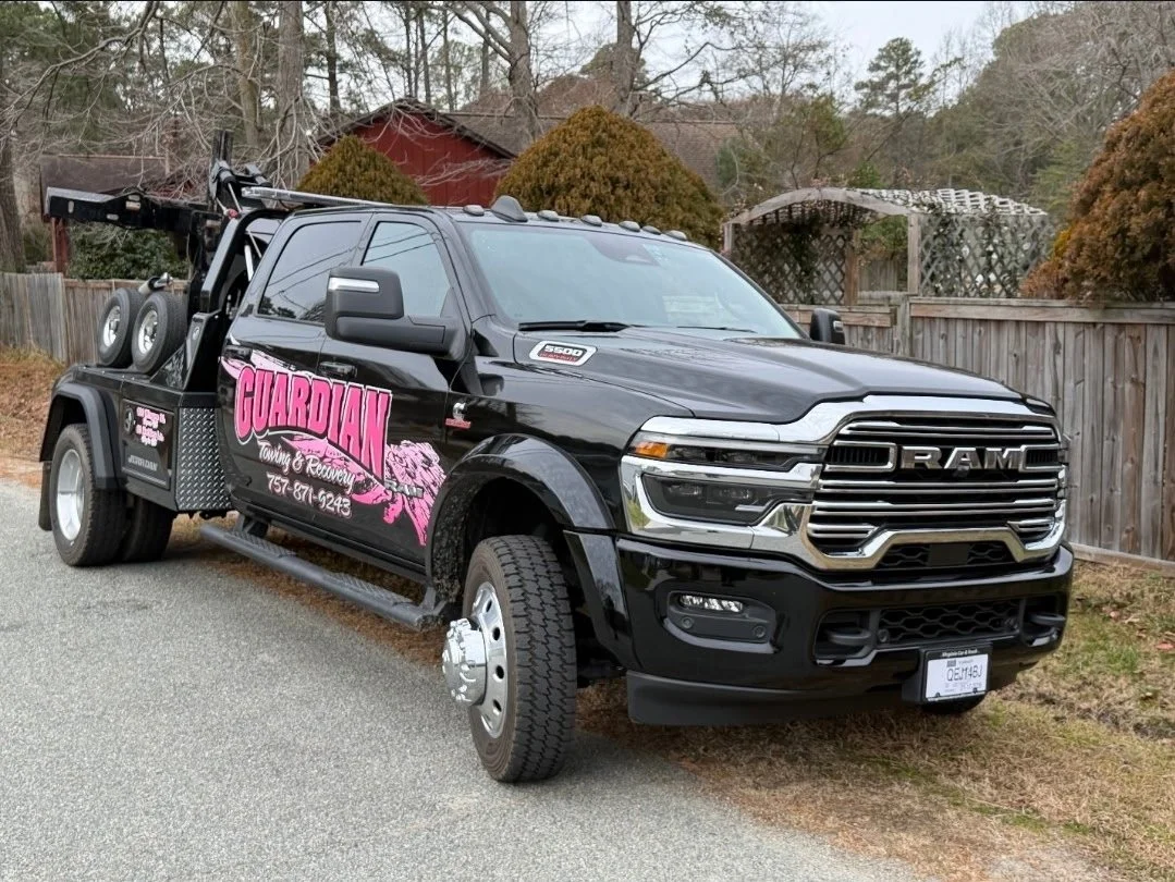 A black RAM tow truck with pink and white Guardian towing and recovery company logo parked on a street with trees and fences in the background.