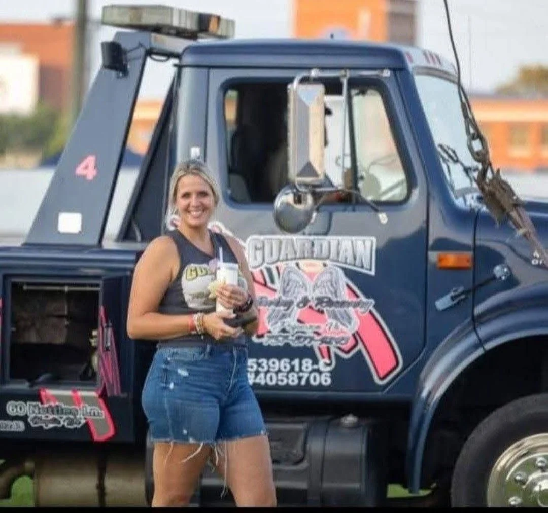 A woman standing in front of a large blue tow truck marked with the word "Guardian" and a logo. She is smiling, holding a drink and a cellphone, wearing a sleeveless top and denim shorts.