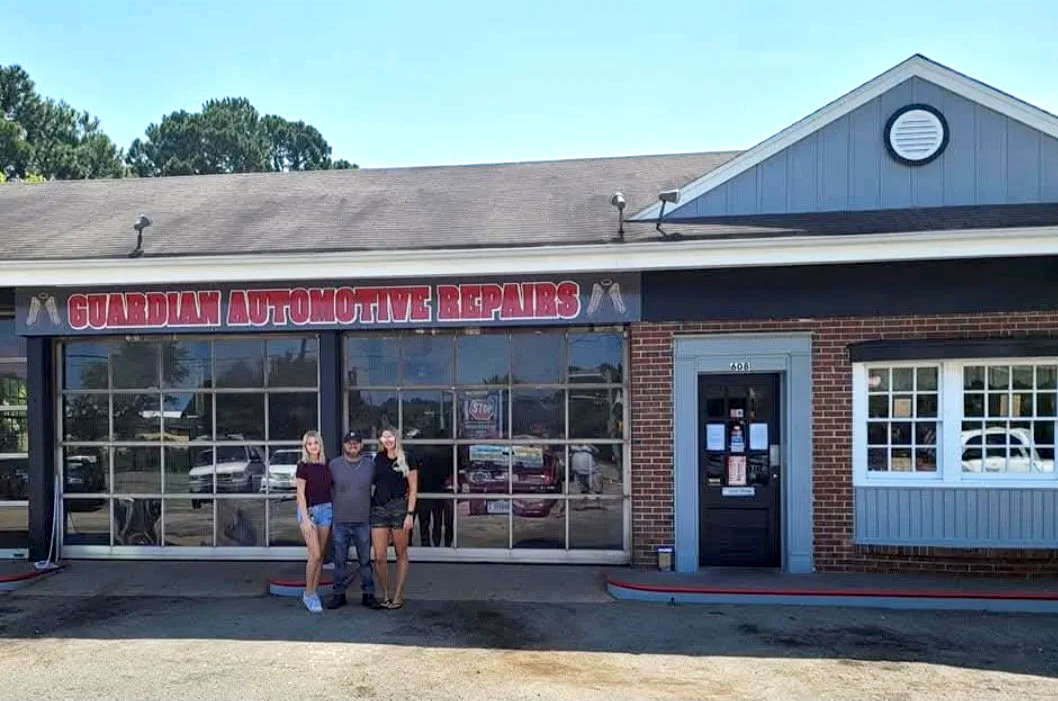 Three people standing in front of Guardian Automotive Repairs shop with large glass windows, brick walls, and a black door, under a sign.