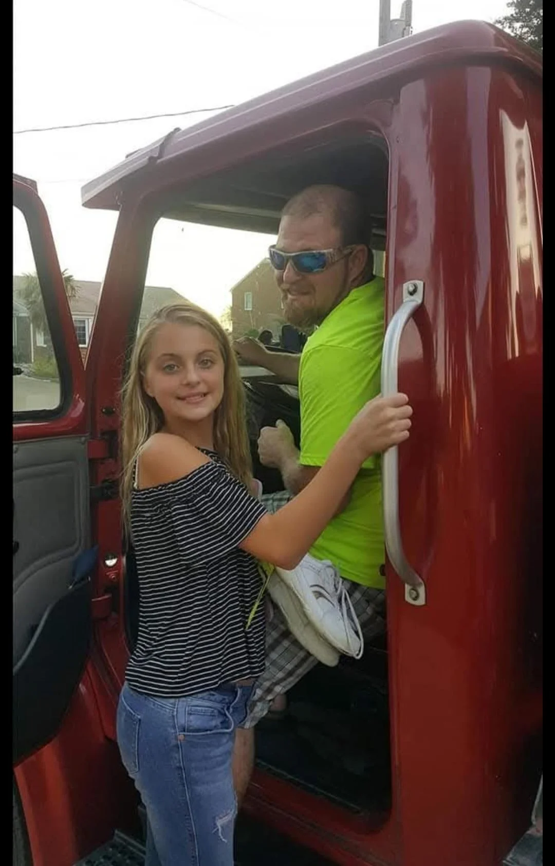 A girl and an adult man are standing inside a red truck, with the man sitting in the driver's seat and the girl standing in the doorway, holding the door handle. The girl is smiling and wearing a striped off-shoulder top and ripped jeans, while the man is wearing sunglasses and a bright yellow shirt, and has a beard. They are outdoors in a residential area.