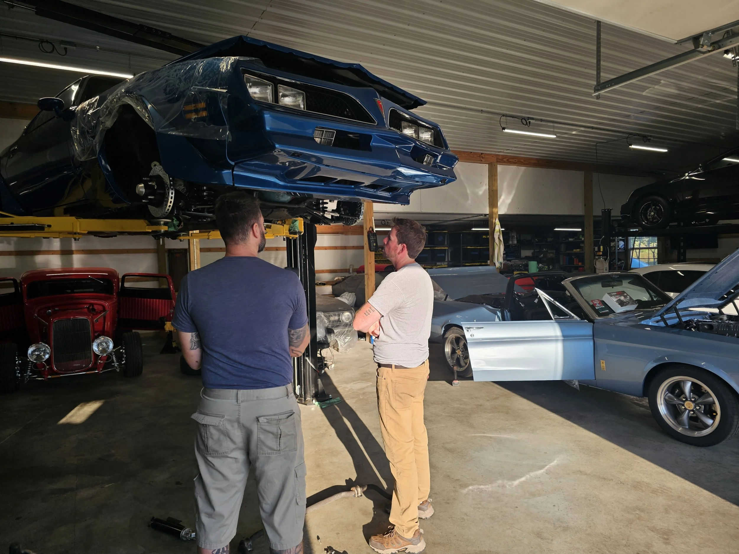 Two men look at a blue race car elevated on a hydraulic lift in a garage filled with various vintage and sports cars.