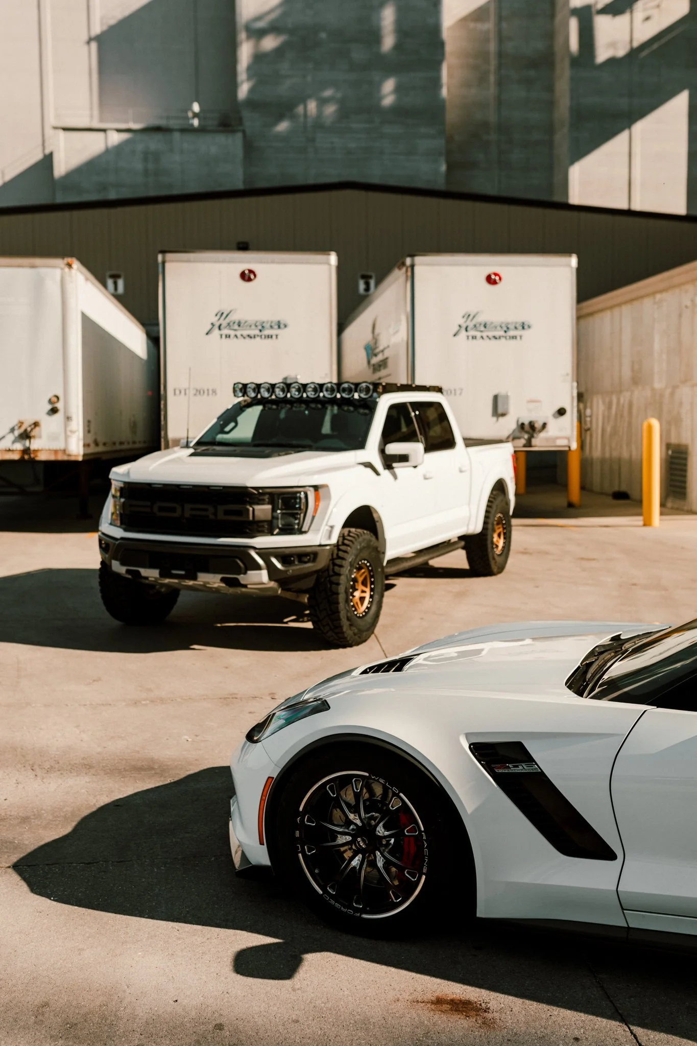 A white pickup truck with off-road tires and a light bar parked in front of storage units, with part of a gray sports car visible in the foreground.