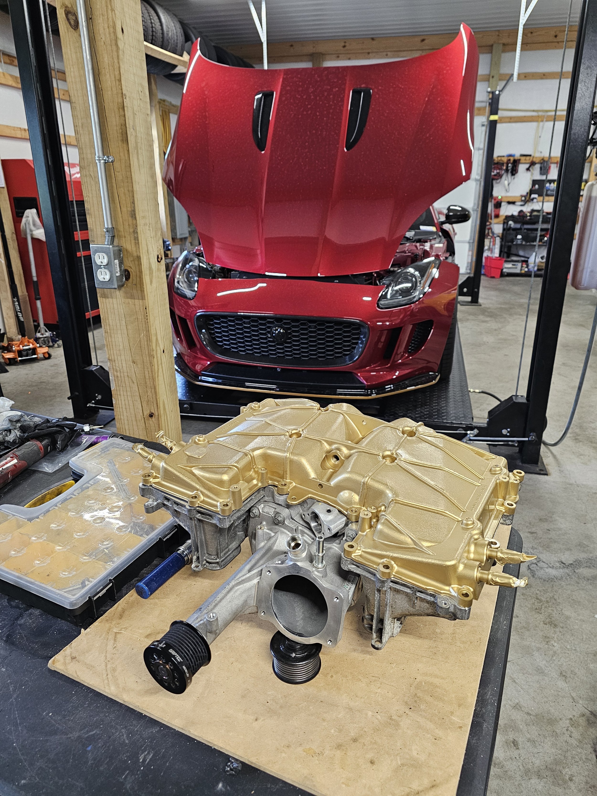 A red sports car with its hood open, situated on a hydraulic lift in a garage. In the foreground, there is an engine or transmission component with a gold-colored top on a workbench.