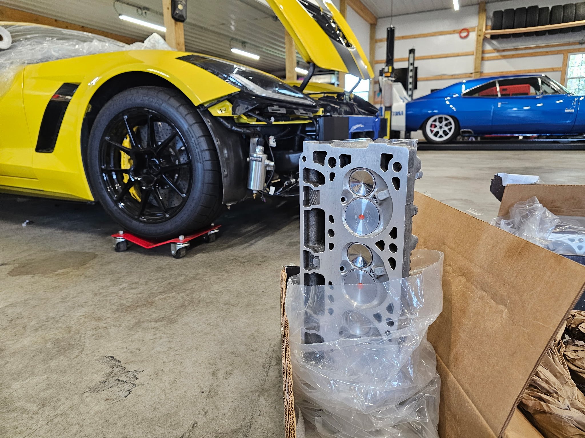 A yellow sports car undergoing repairs on a jack in a garage, with an engine part and a blue classic car in the background.
