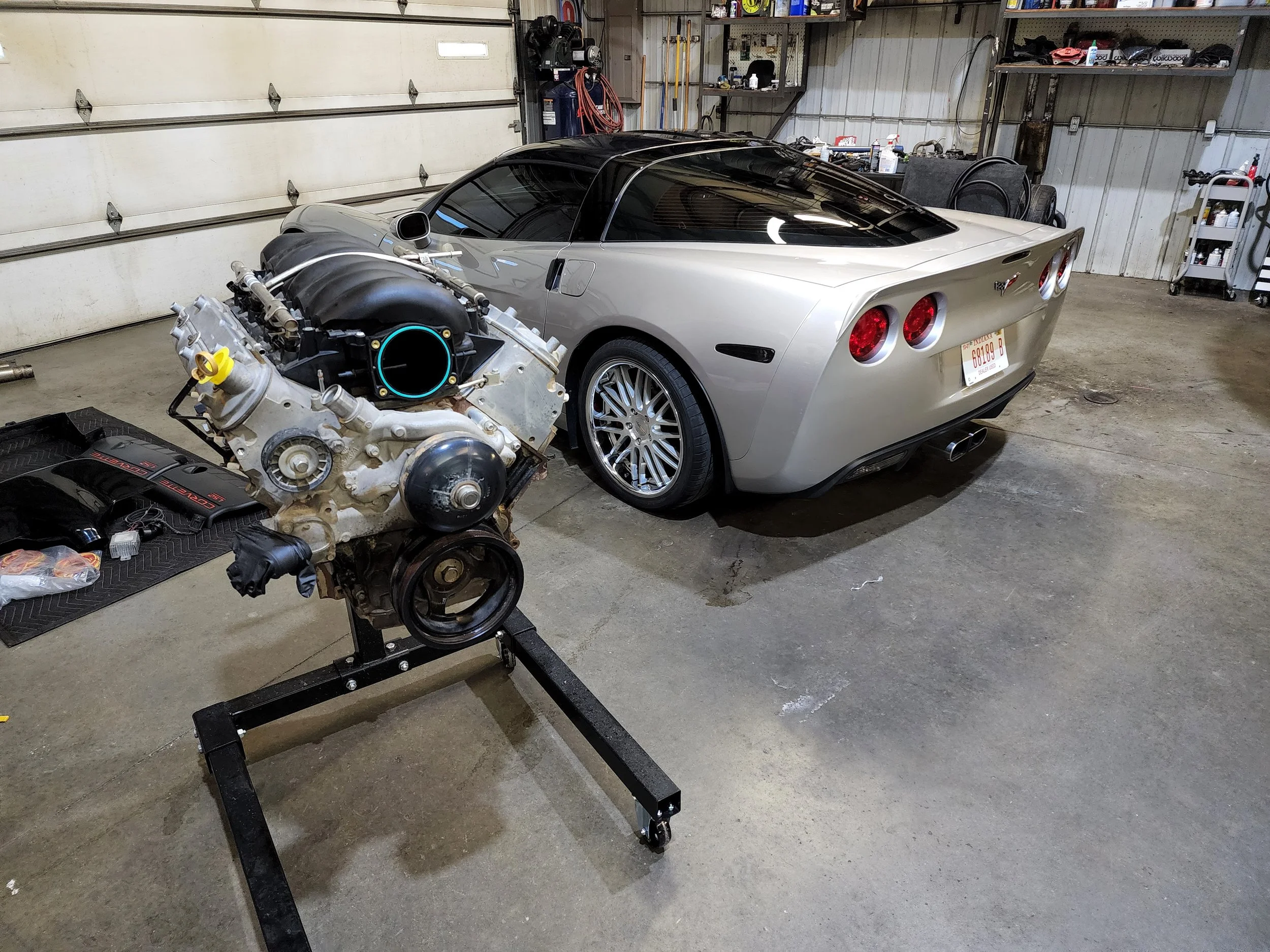 A silver Chevrolet Corvette with a black roof parked inside a garage. An engine is mounted on a stand nearby.