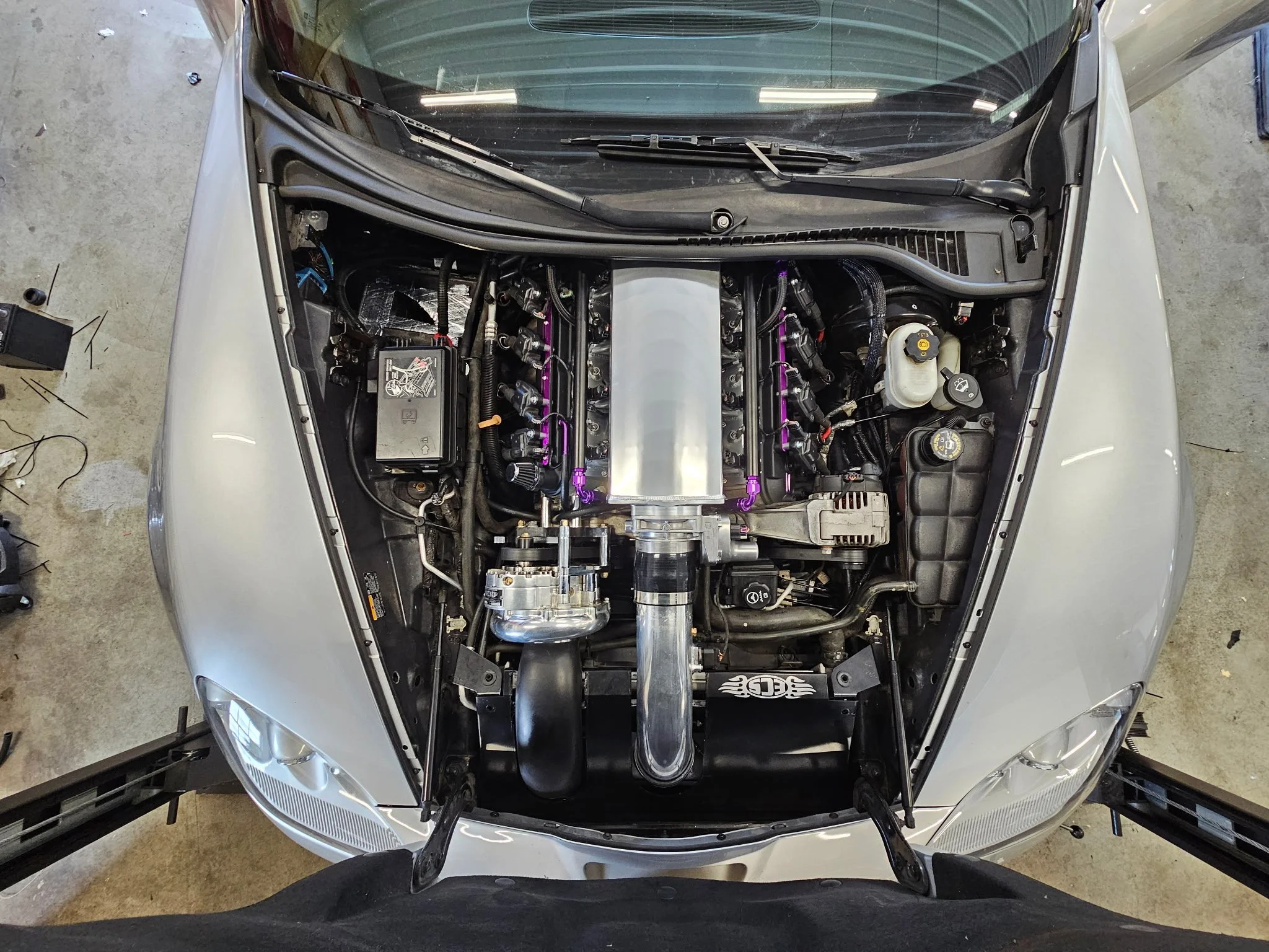 Inside view of a sports car engine bay, showing performance modifications including a large central aluminum intake manifold, custom piping, and various engine components.